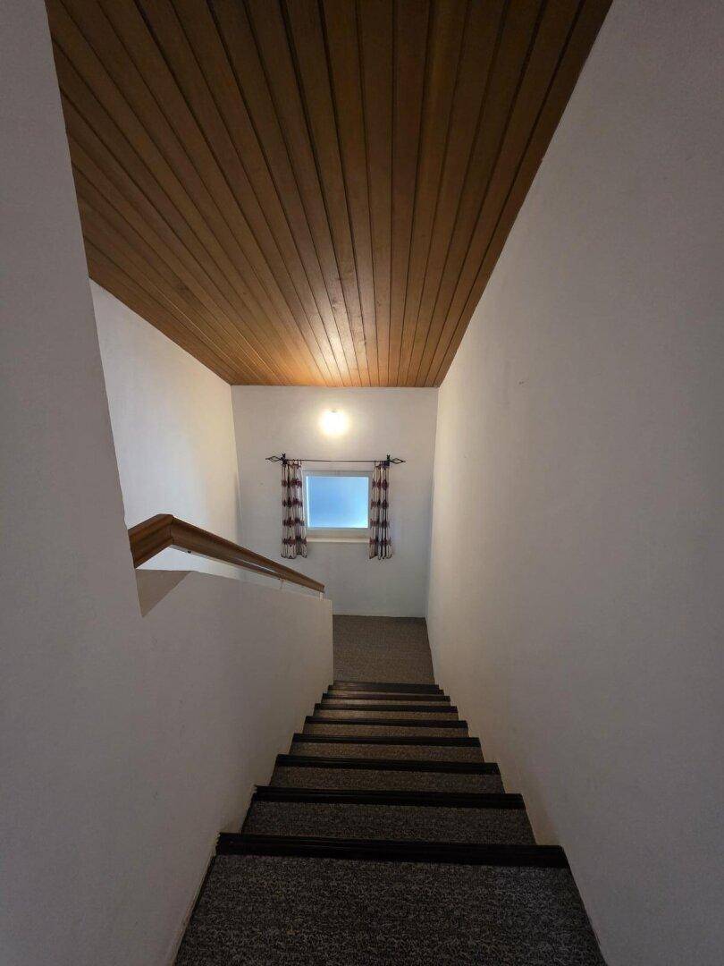 Treppenhaus mit Teppichstufen, weißen Wänden und einer Holzdecke, hell durch ein kleines Fenster.