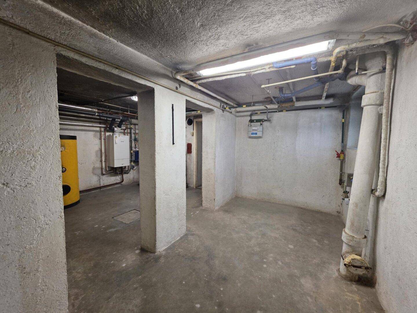 Kellerraum mit Betonboden, sichtbaren Rohren und technischen Installationen.