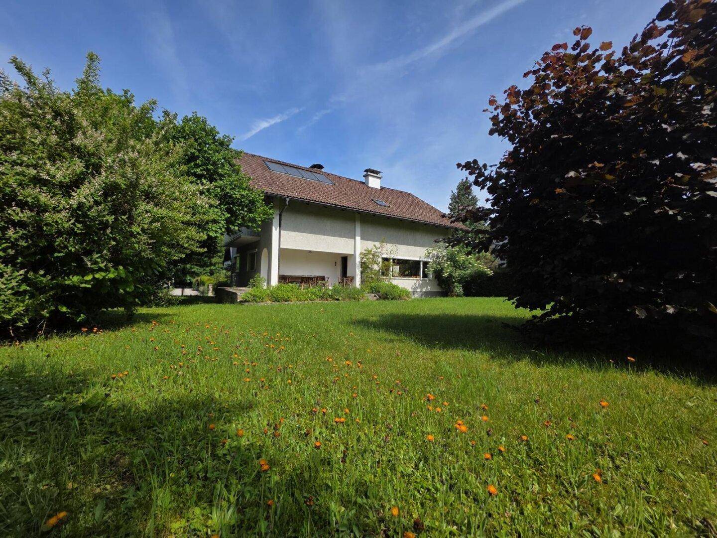 Ansicht des Hauses von der großen Rasenfläche aus, umgeben von Bäumen und viel Grün.