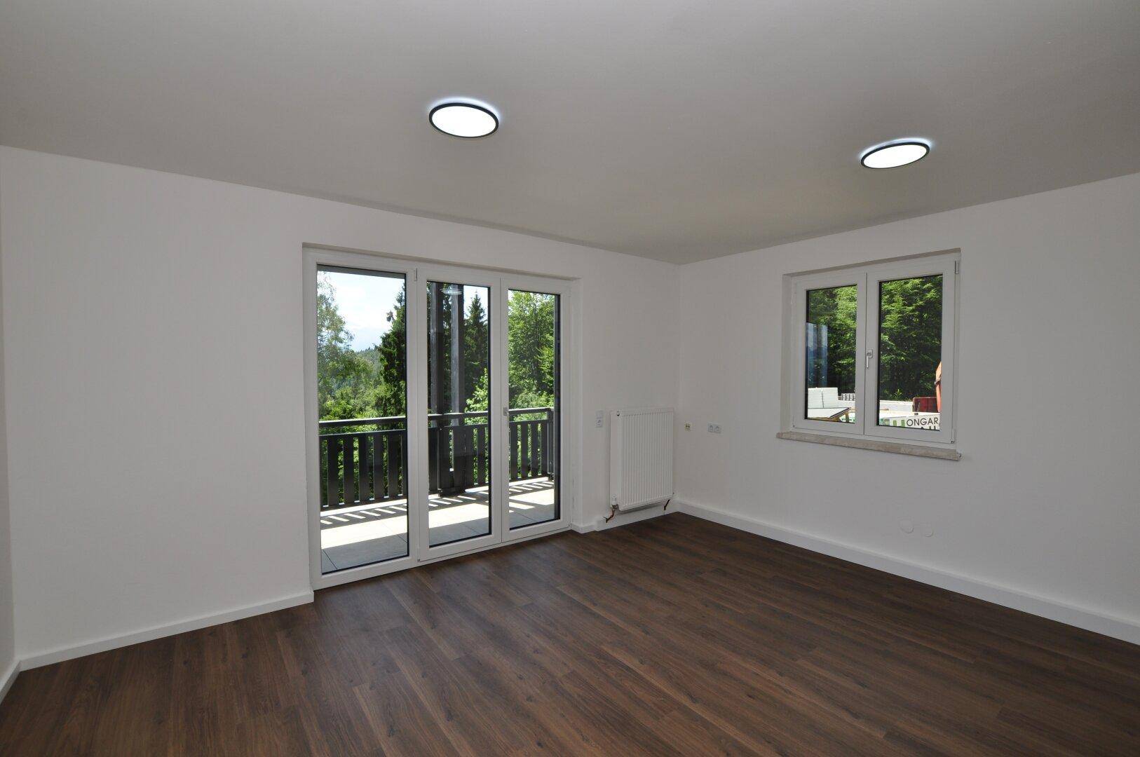 Helles, leeres Zimmer mit dunklem Holzboden, weißen Wänden und Zugang zu einem Balkon mit Waldblick.