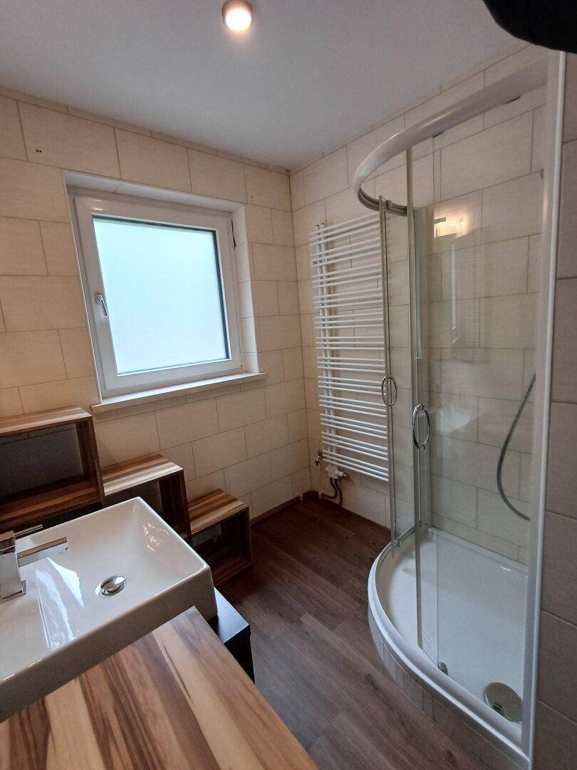 Helles Badezimmer mit Duschkabine, beigefarbenen Fliesen und einem Fenster für natürliche Belüftung.