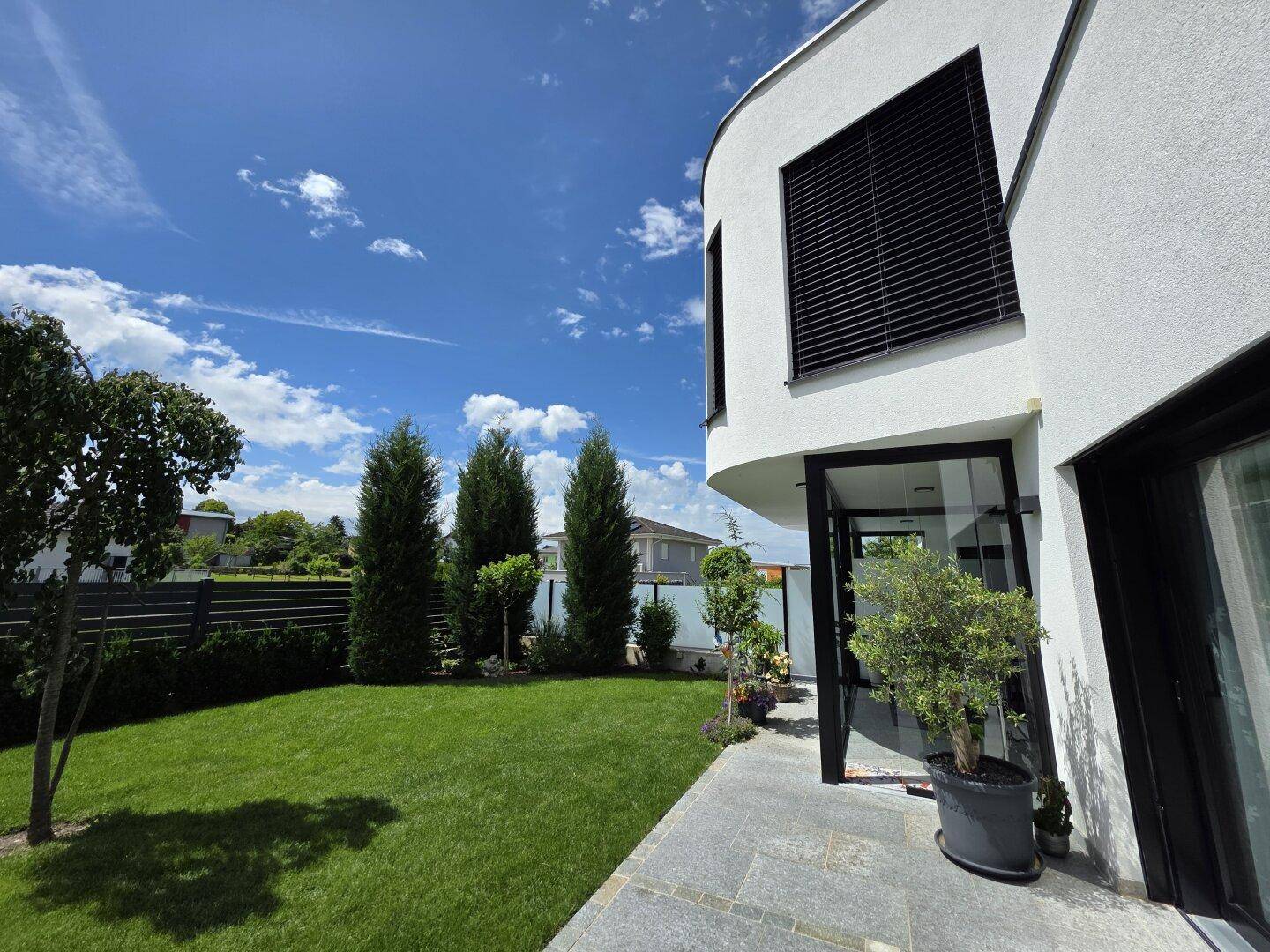 Seitlicher Blick auf die moderne Villa mit großen Fenstern, Außenjalousien und einem grünen Garten.