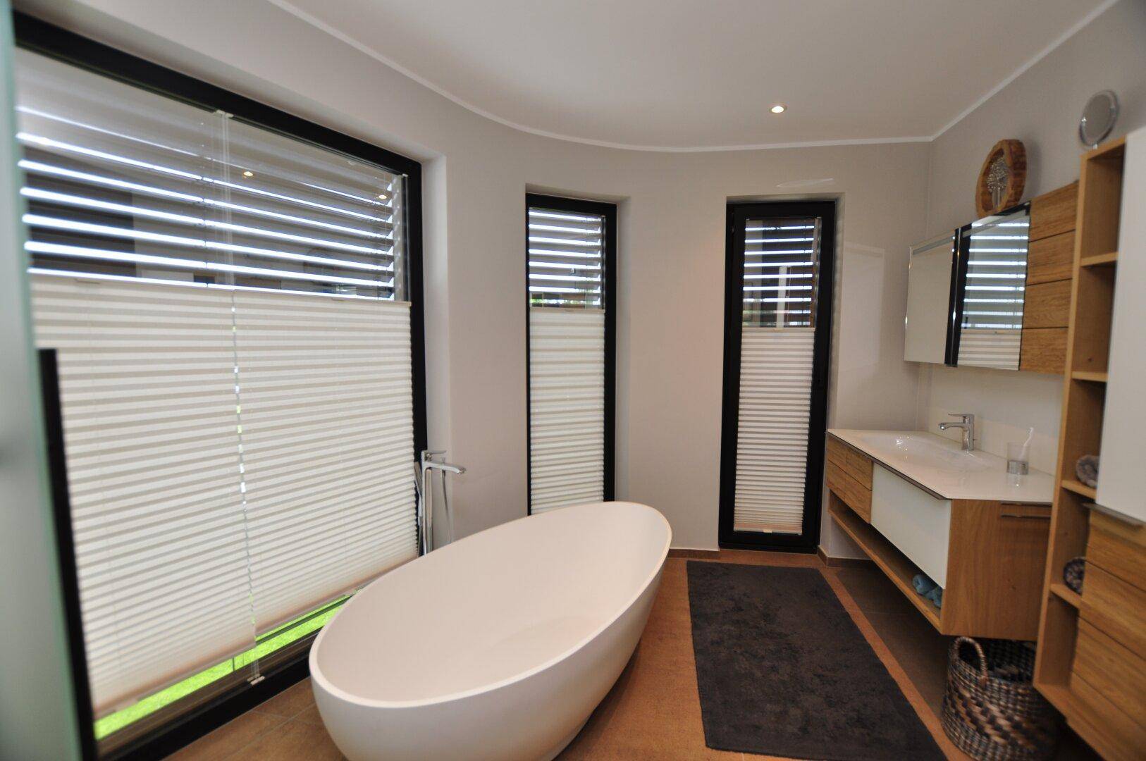 Modernes Badezimmer mit freistehender Badewanne, Holzwaschtisch und großen Fenstern mit Plissees.