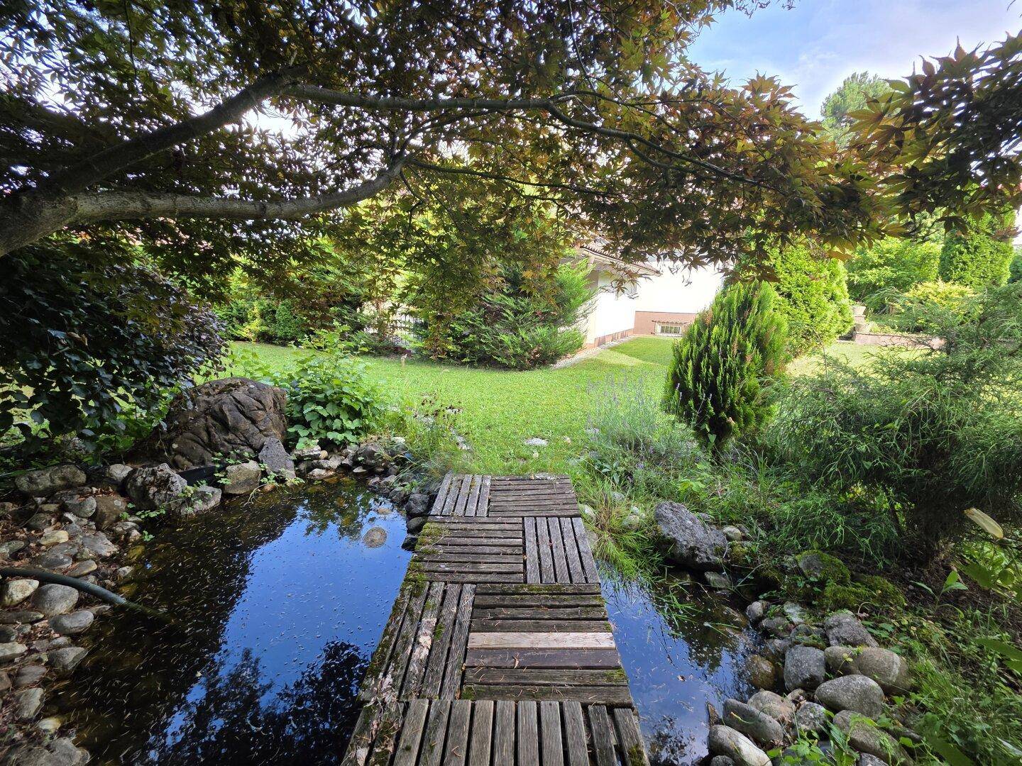 Idyllischer Gartenteich mit kleiner Holzbrücke, umgeben von üppiger Vegetation und schattenspendenden Bäumen.