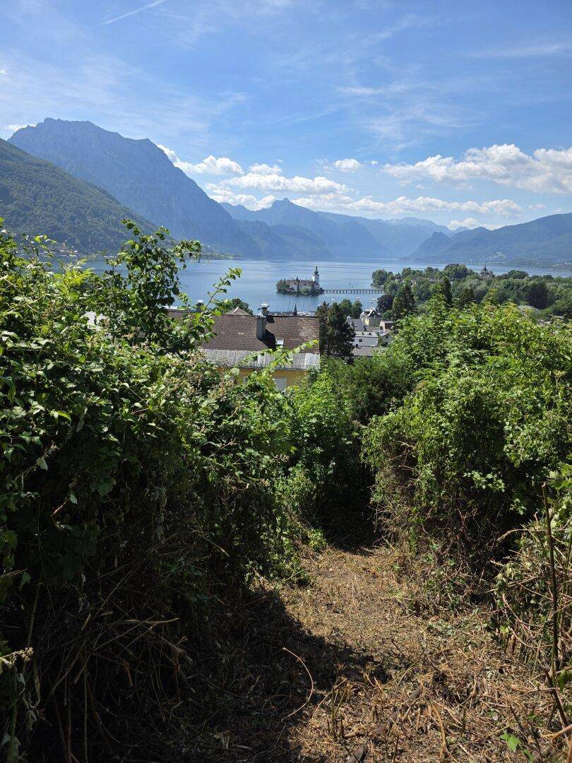 Schmaler Pfad durch dichtes Grün, der sich zu einem weiten Blick auf See und Berge öffnet.