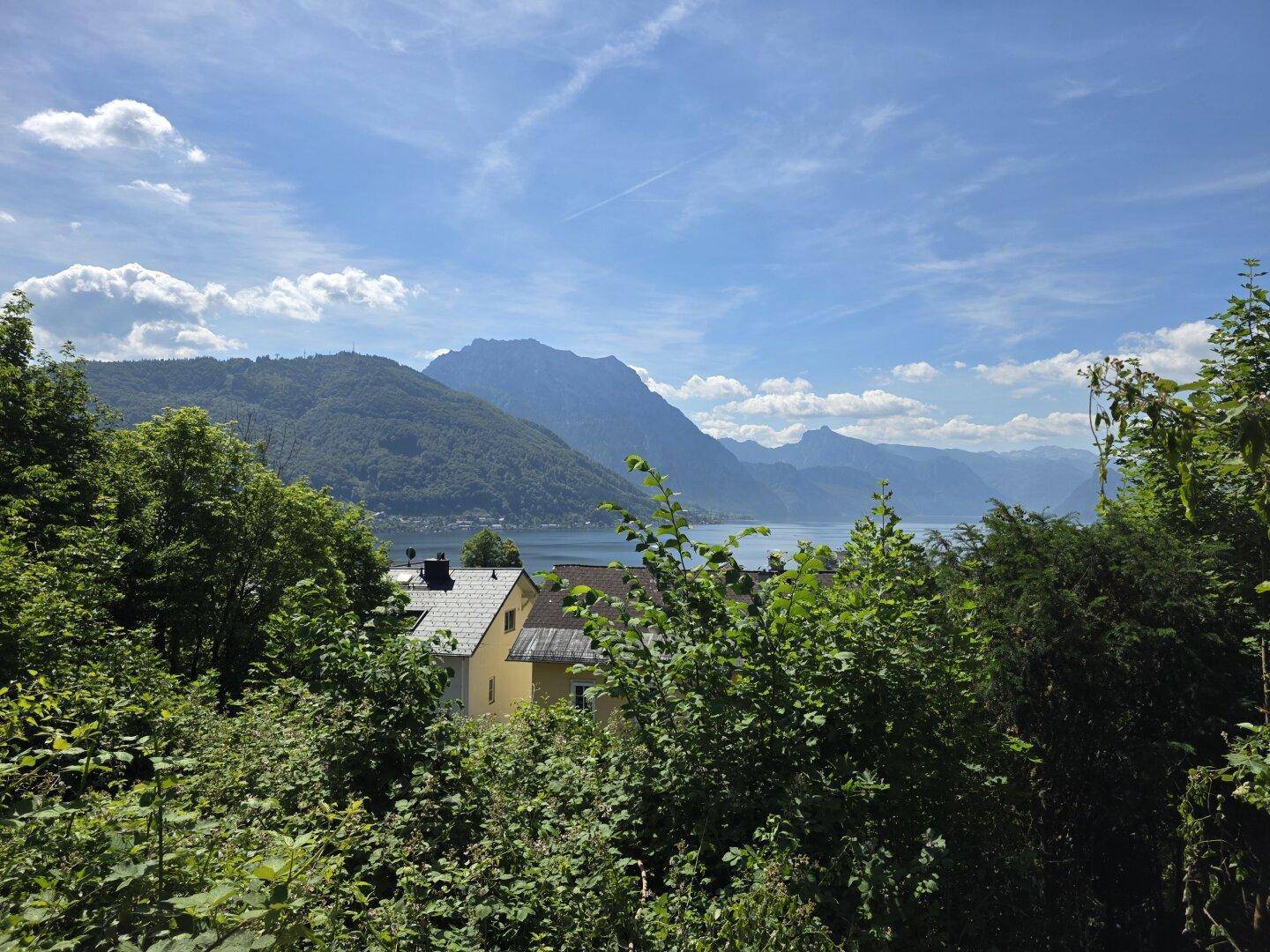 Blick über grüne Bäume und Sträucher auf einen See und Berge in der Ferne, mit einem Haus im Vordergrund.