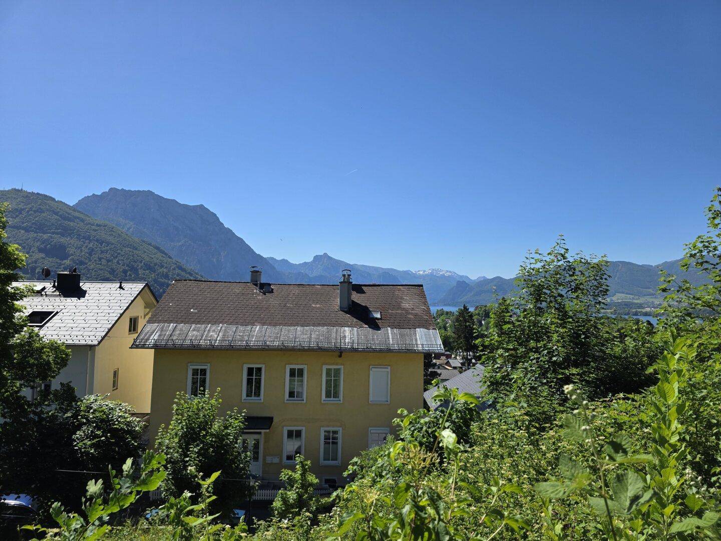 Gelbes Haus mit dunklem Dach, umgeben von üppigem Grün und mit Bergblick auf einen See.