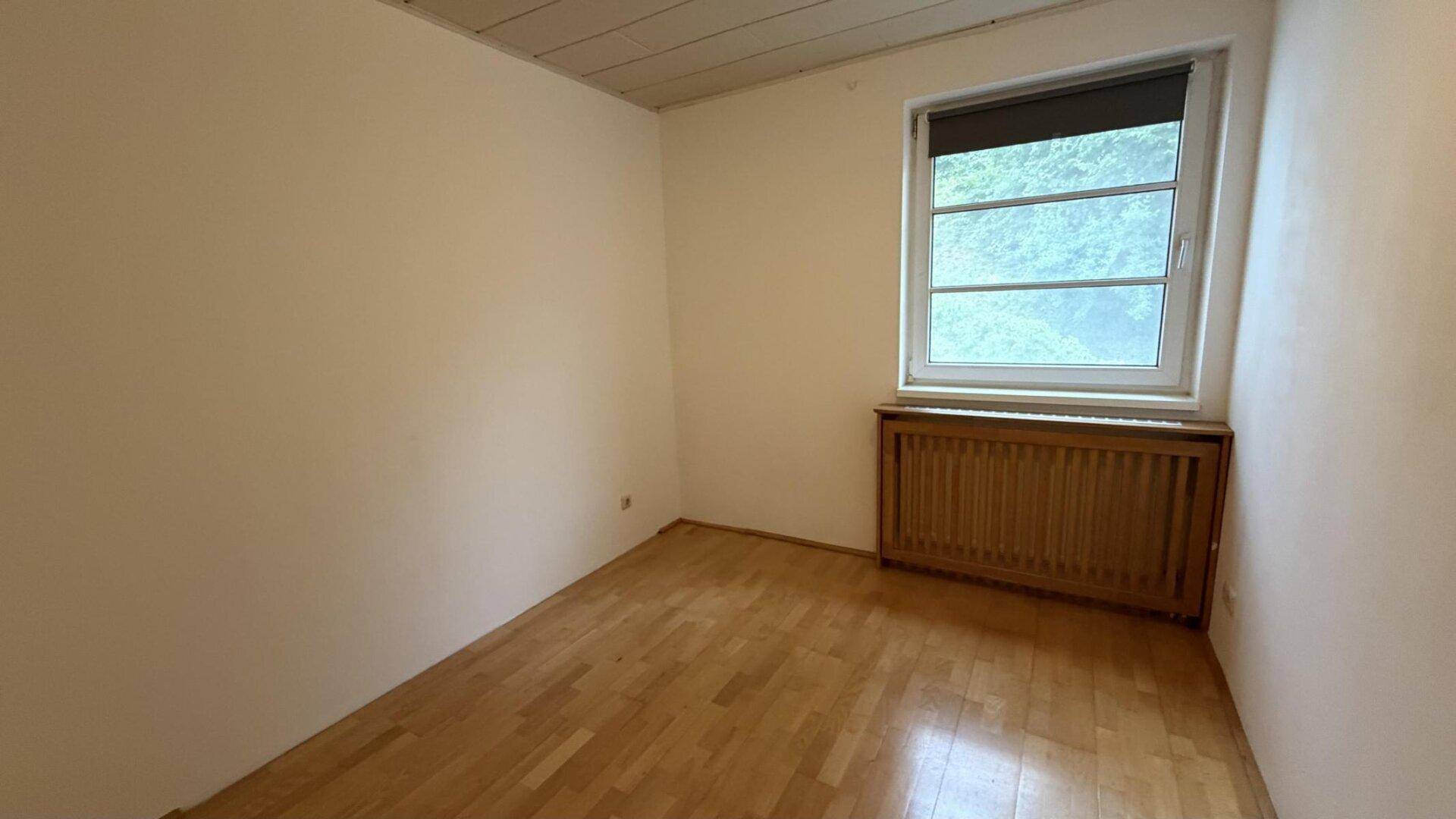 Helles Schlafzimmer mit Parkettboden, weißen Wänden und Fenster mit Heizkörperverkleidung.