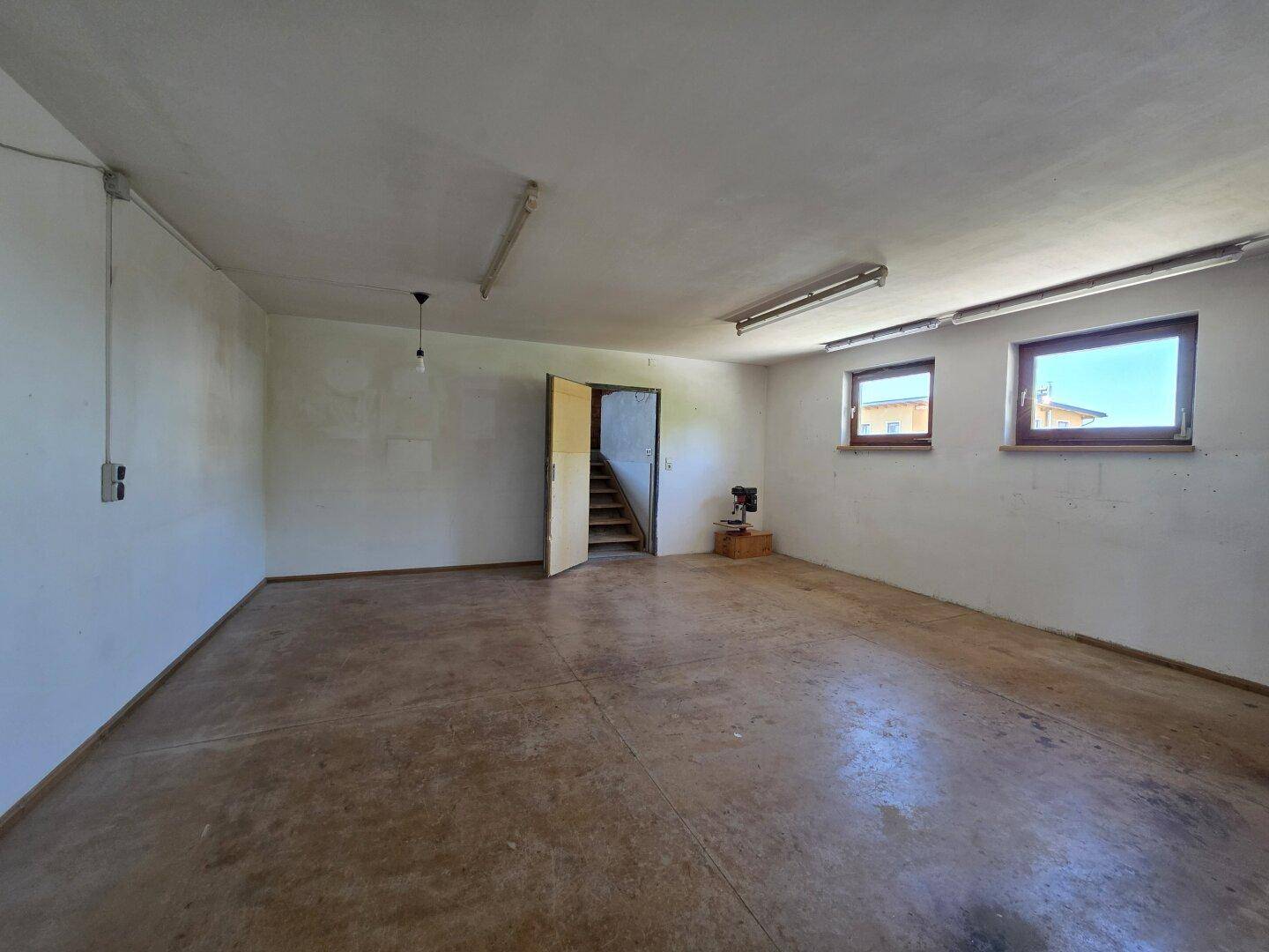 Großer leerer Raum mit Betonboden und zwei Fenstern, ideal als Werkstatt oder Lager.
