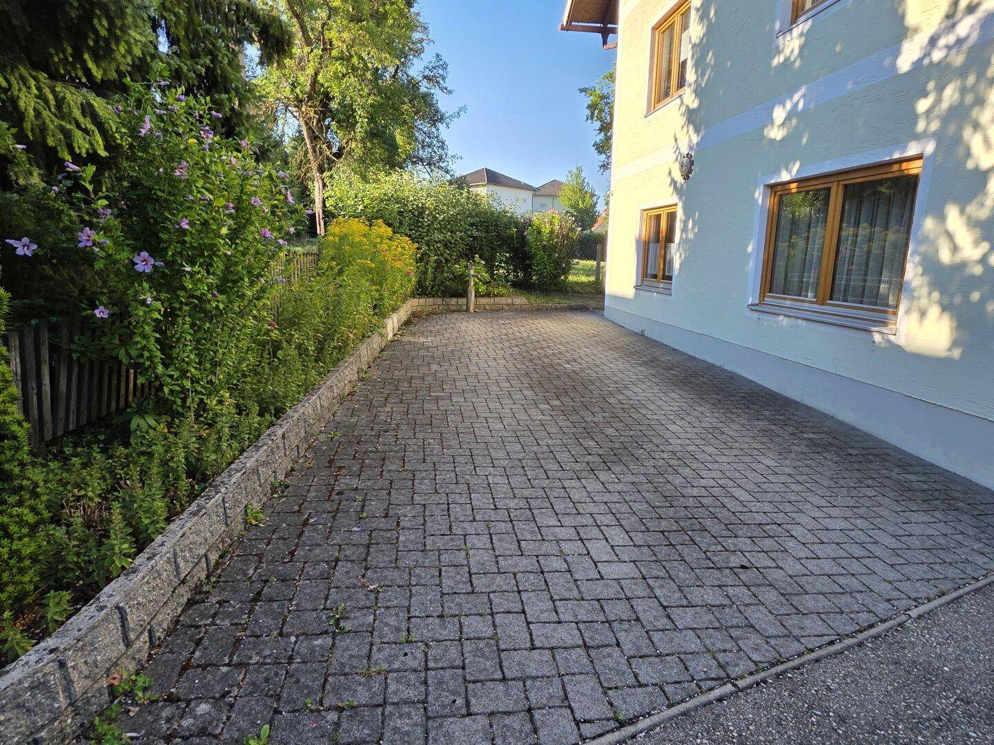 Gepflasterte Einfahrt neben dem Haus mit grüner Bepflanzung und Holzzaun.