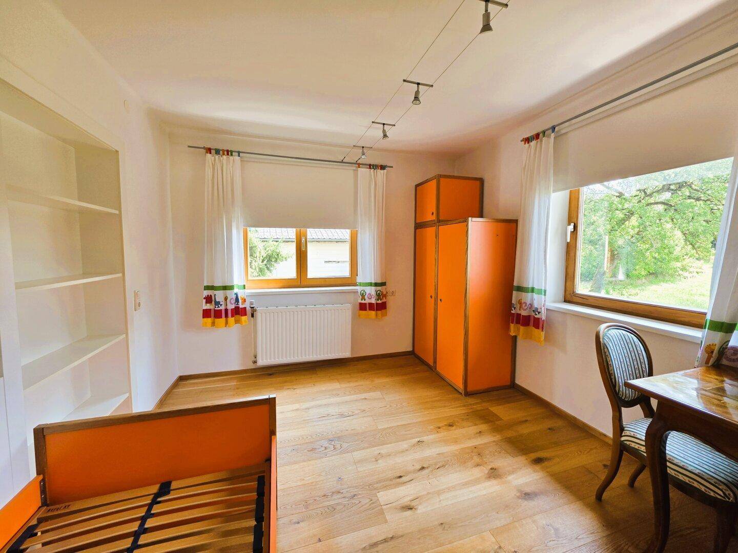 Kinderzimmer mit Holzboden, orangefarbenen Möbeln und Blick ins Grüne.