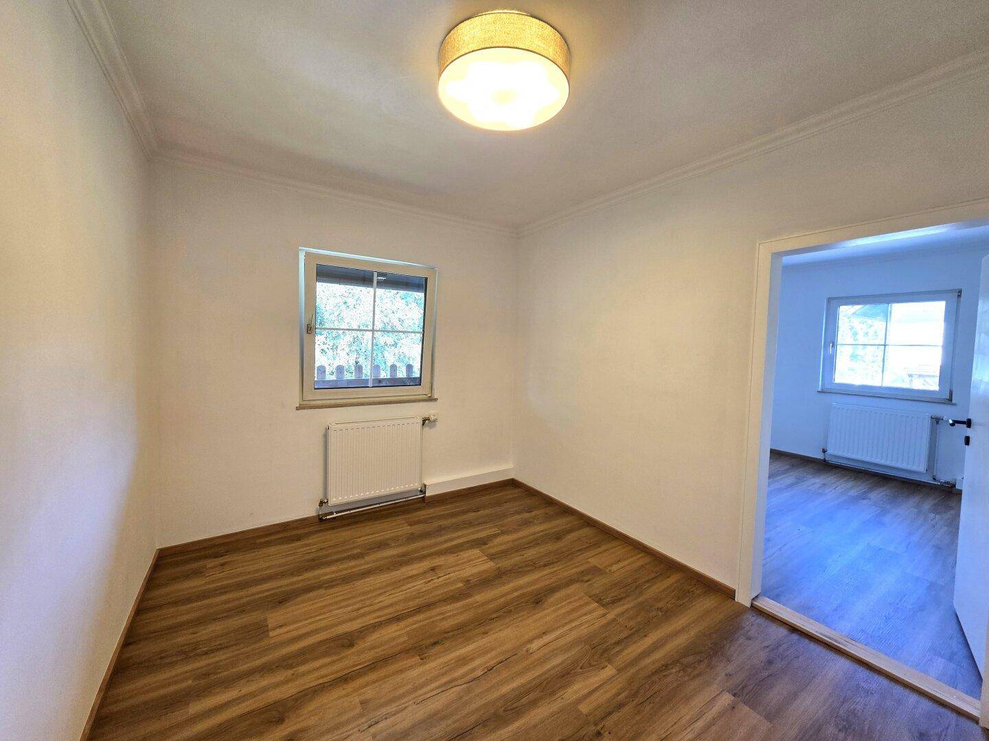 Helles Schlafzimmer mit Laminatboden, Heizkörper und Fenster mit Blick ins Grüne.