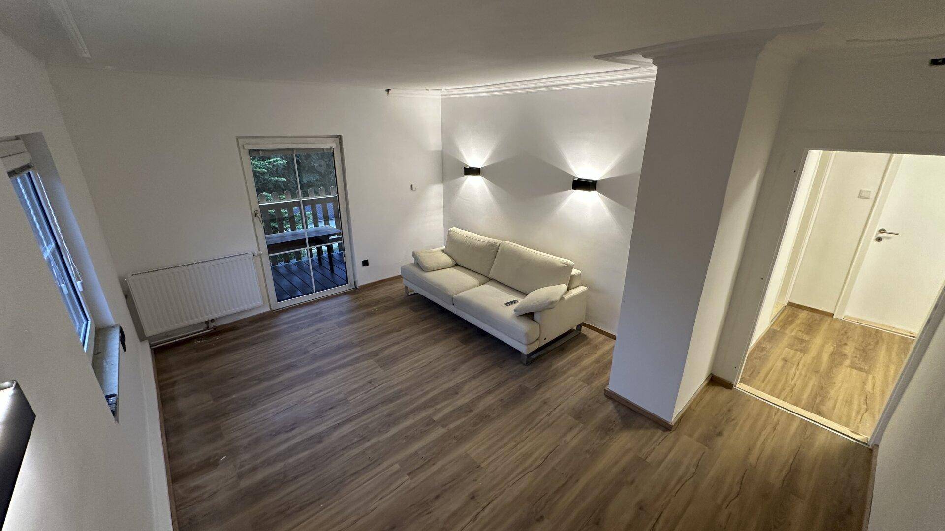 Geräumiges Wohnzimmer mit Balkonzugang, Laminatboden und modernen Wandleuchten.