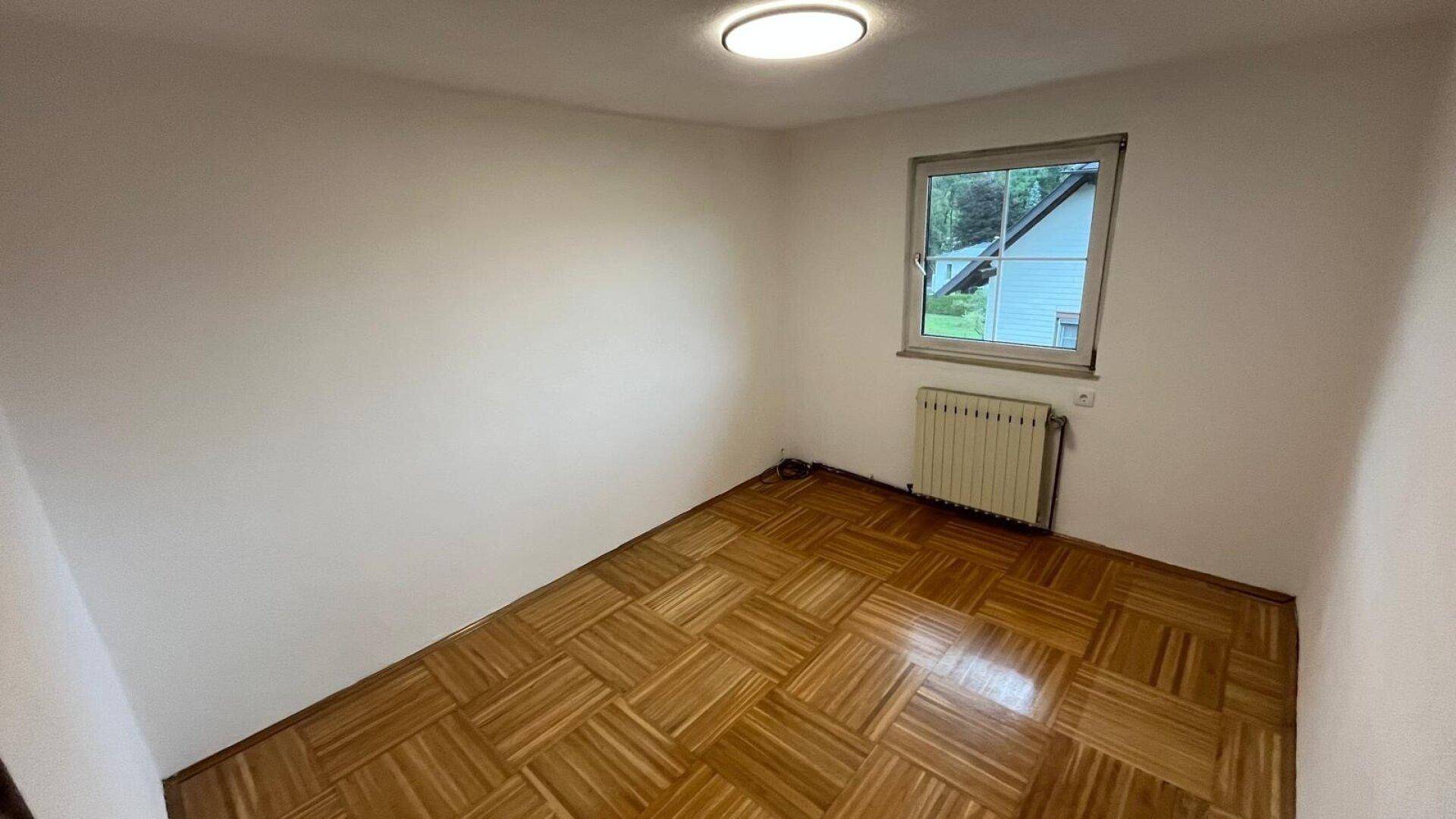 Schlafzimmer mit Parkettboden, Heizkörper und Fenster mit Blick ins Grüne.