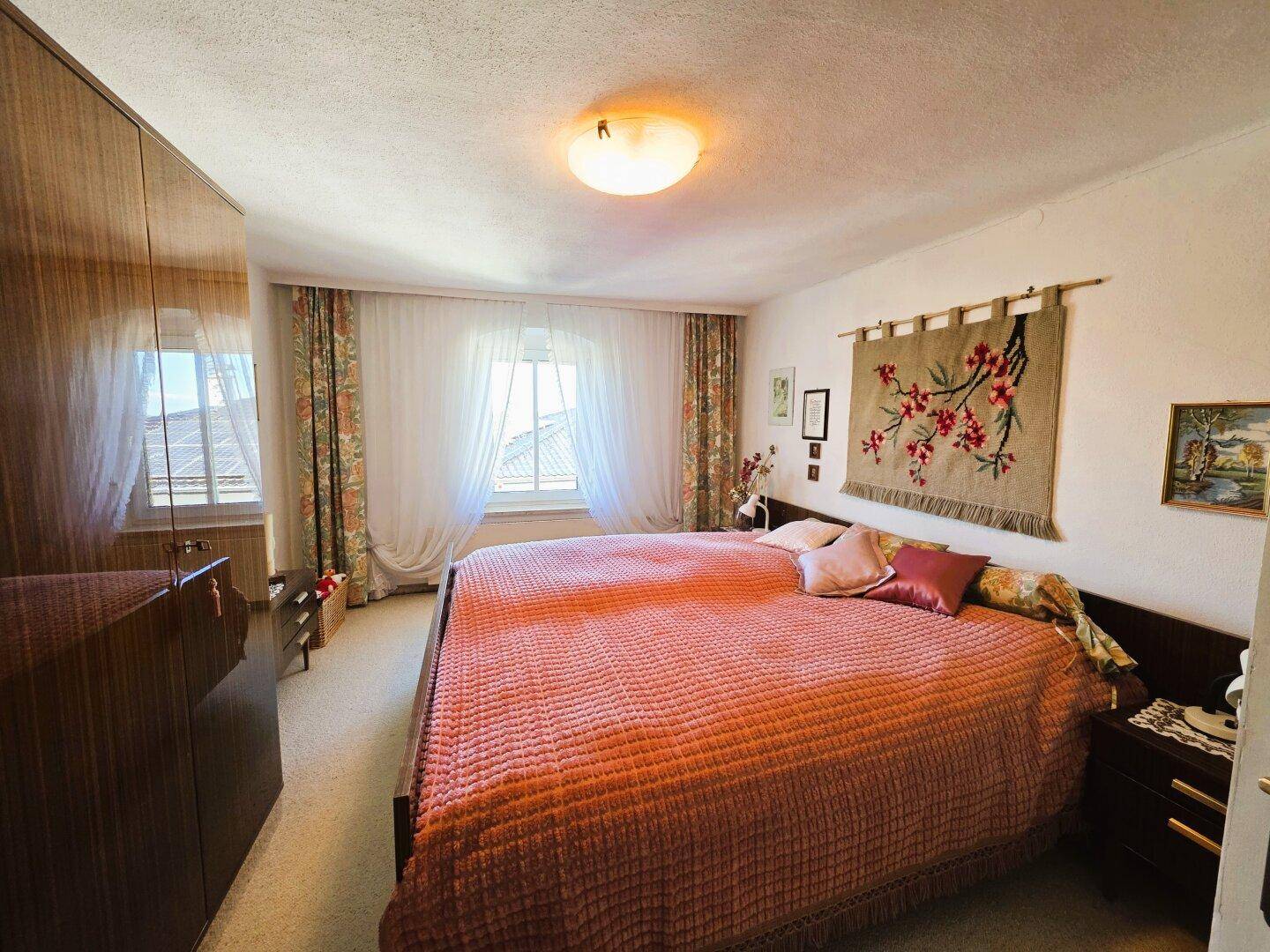 Helles Schlafzimmer mit Doppelbett, Holzschrank und Fenstern mit gemusterten Vorhängen.