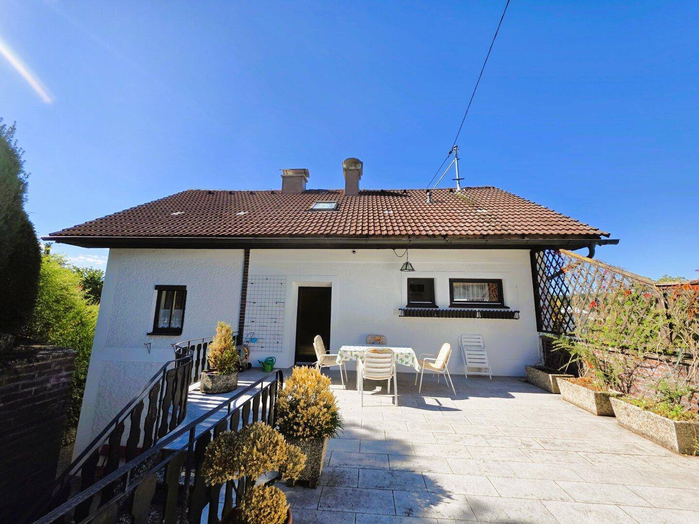 Rückseite des Hauses mit einer großzügigen, gepflasterten Terrasse und Gartenmöbeln unter blauem Himmel.