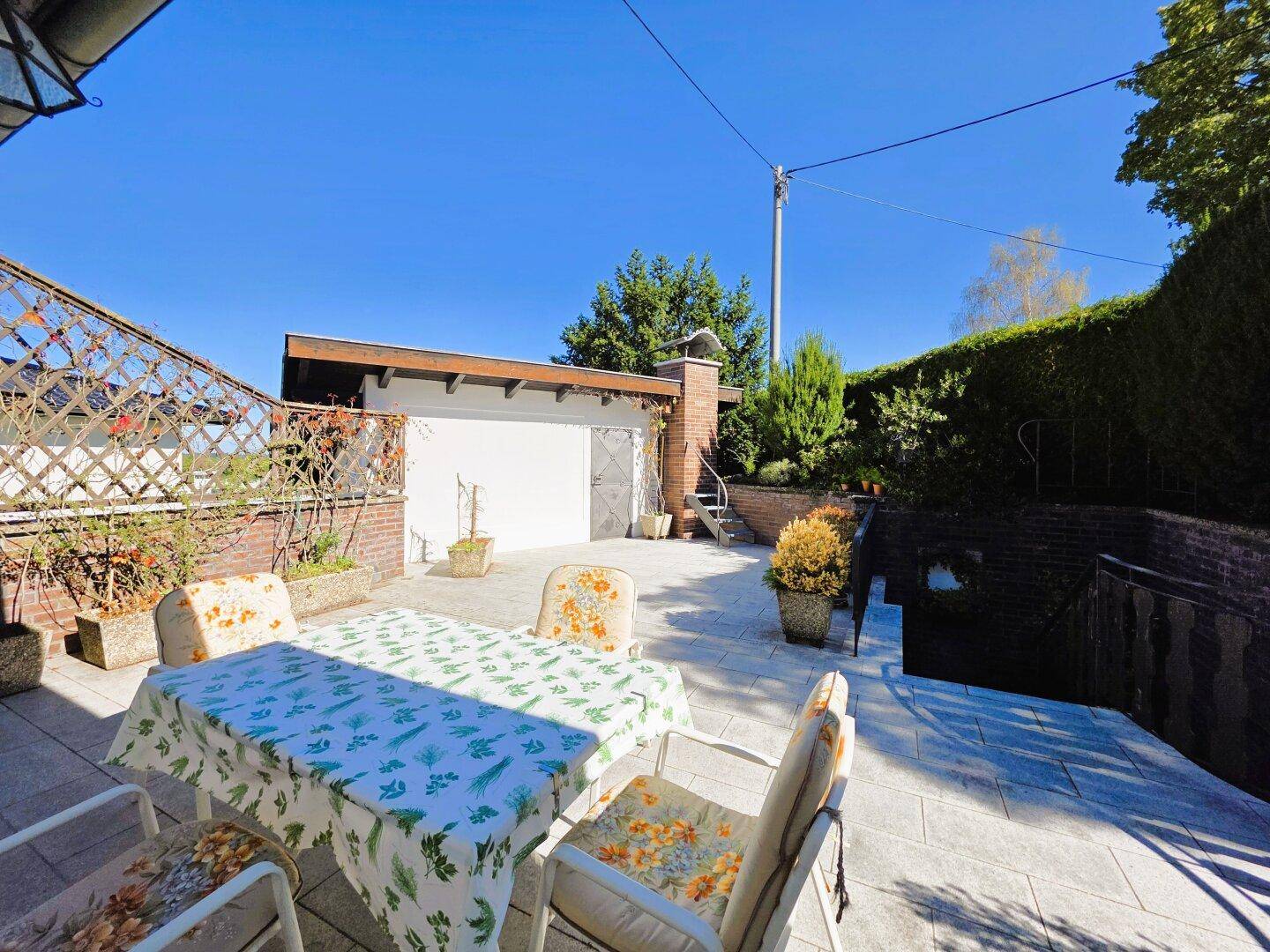 Weitläufige Terrasse mit Esstisch und Stühlen, angrenzend an eine Garage und Treppenabgang.