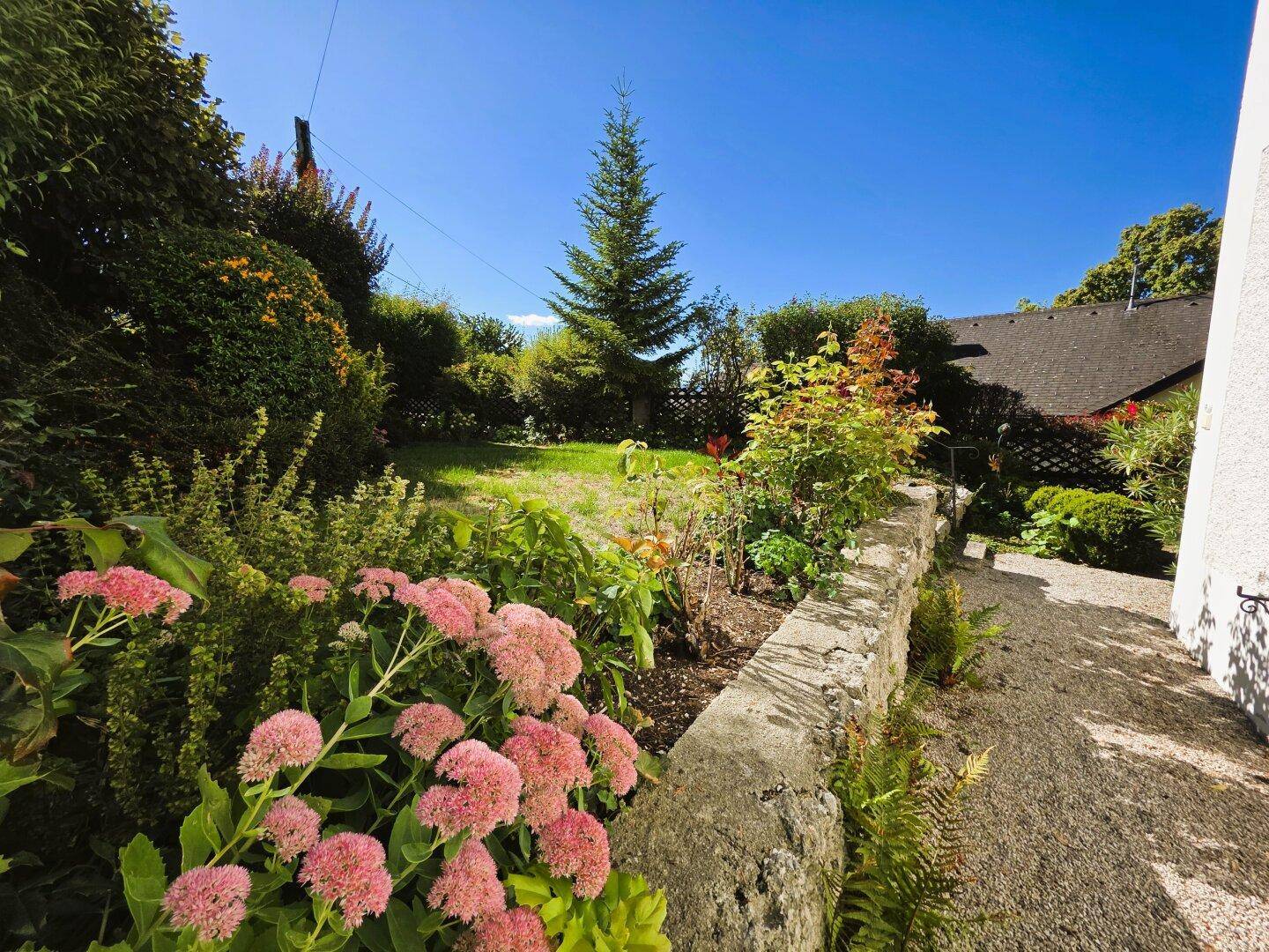Gepflegter Gartenbereich mit blühenden Pflanzen, einer Steinmauer und einer Rasenfläche unter blauem Himmel.