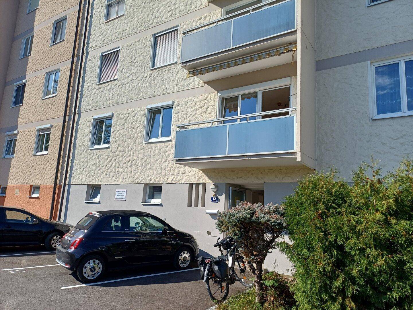 Fassade eines Wohngebäudes mit mehreren Balkonen und geparkten Autos auf dem dazugehörigen Parkplatz.