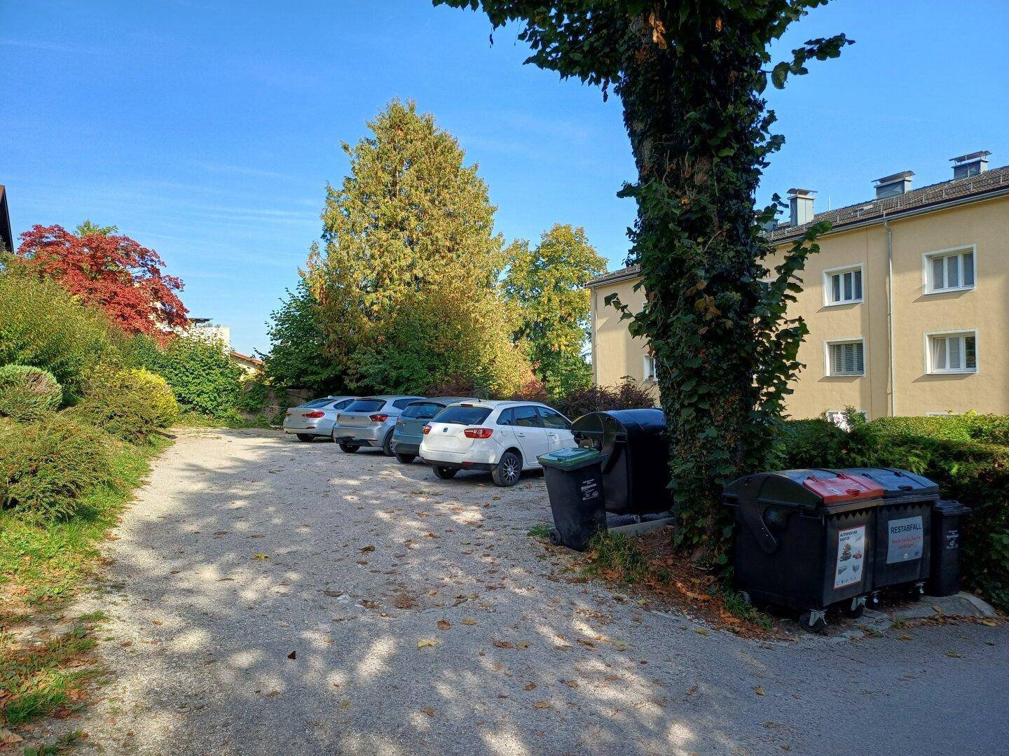 Außenparkplatz mit mehreren Autos und Bäumen neben einem Wohngebäude, Mülltonnen sichtbar.