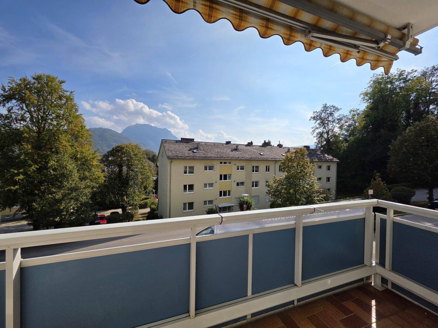 Blick vom Balkon mit Markise auf ein Wohngebäude und die umliegende Berglandschaft unter blauem Himmel.