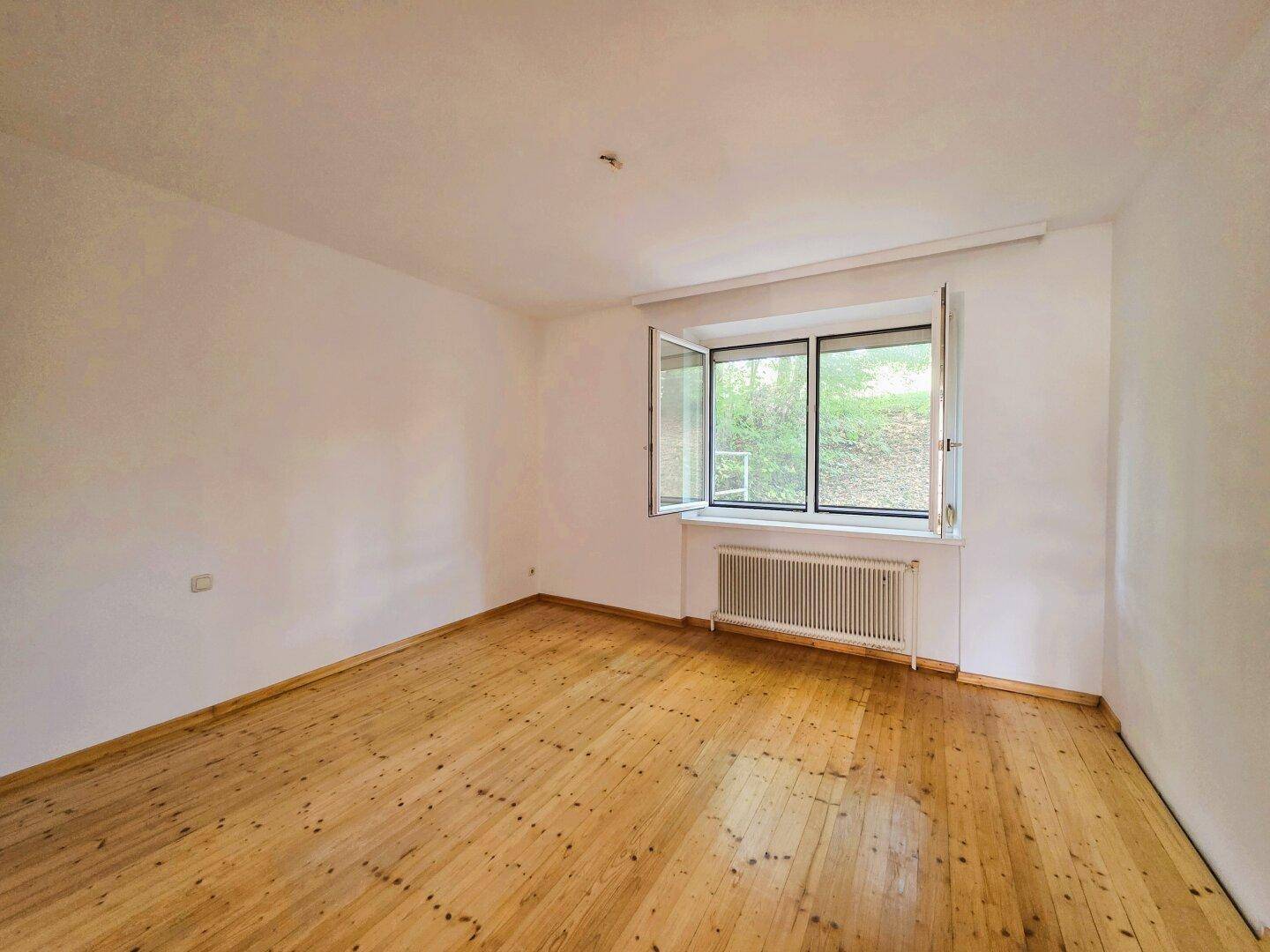 Helles, leeres Zimmer mit Holzdielenboden, weißen Wänden und Blick ins Grüne durch das Fenster.