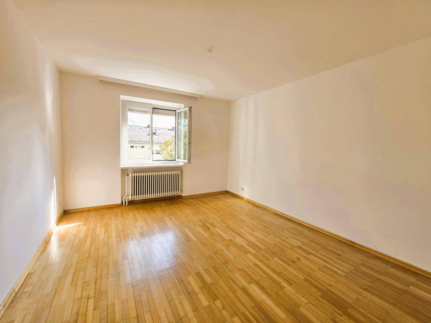 Leeres Zimmer mit Holzboden, weißen Wänden und einem Fenster, das einen Blick auf andere Gebäude bietet.