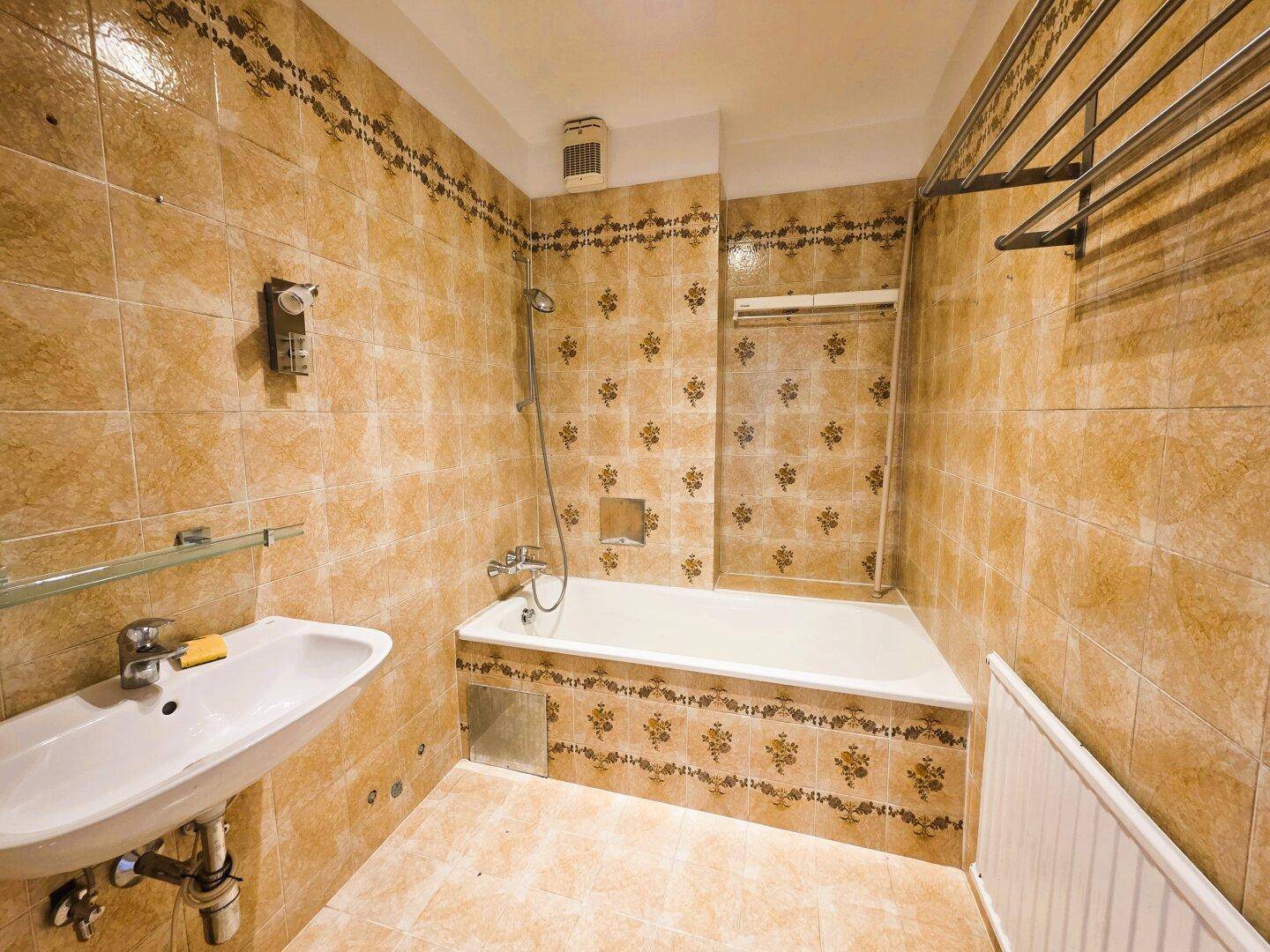 Badezimmer im Stil der 80er Jahre mit gemusterten braunen Fliesen, Badewanne mit Dusche und Waschbecken.