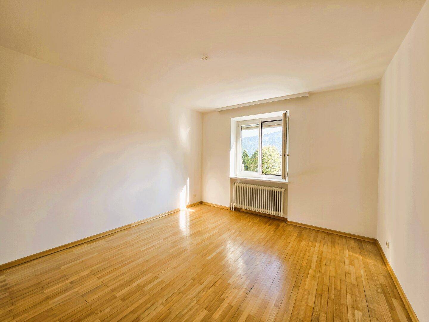Helles, leeres Zimmer mit Parkettboden, weißen Wänden und einem Fenster mit Blick ins Grüne.