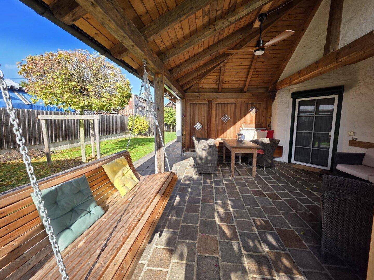 Überdachte Terrasse mit Holzschaukel, Sitzgelegenheiten und Deckenventilator für entspannte Stunden.