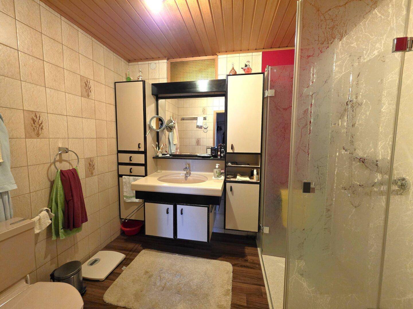 Badezimmer mit Duschkabine, Waschtisch und Holzpaneeldecke im 80er-Jahre-Stil.