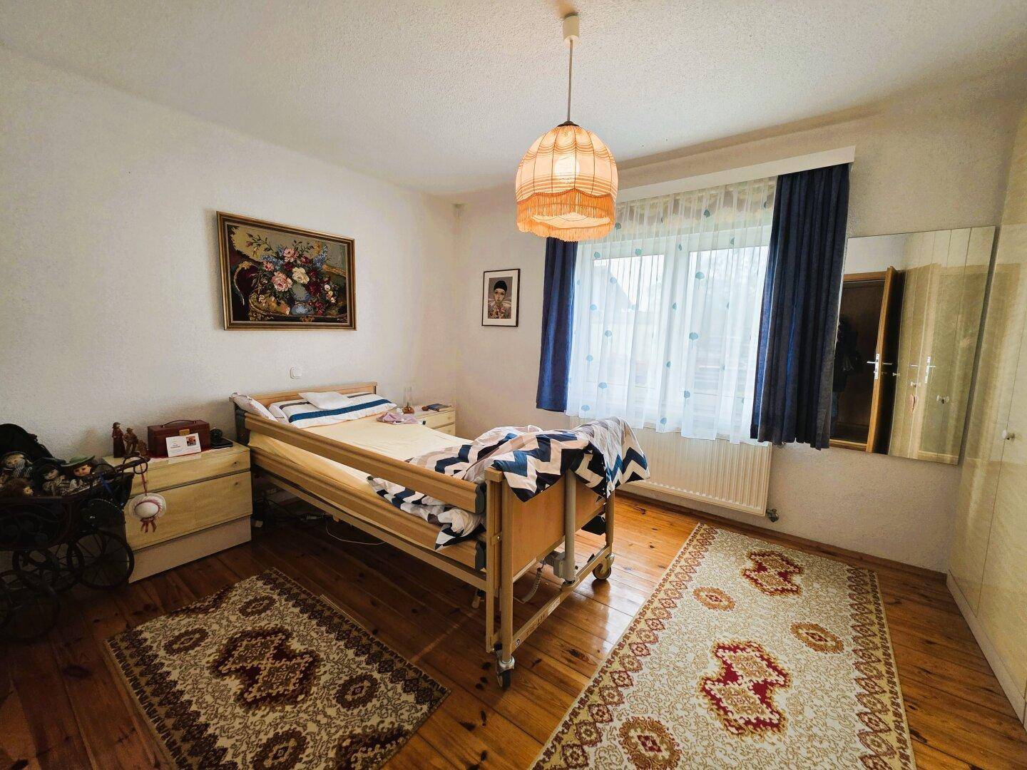 Schlafzimmer mit Pflegebett, Holzparkett und traditionellem Teppich.