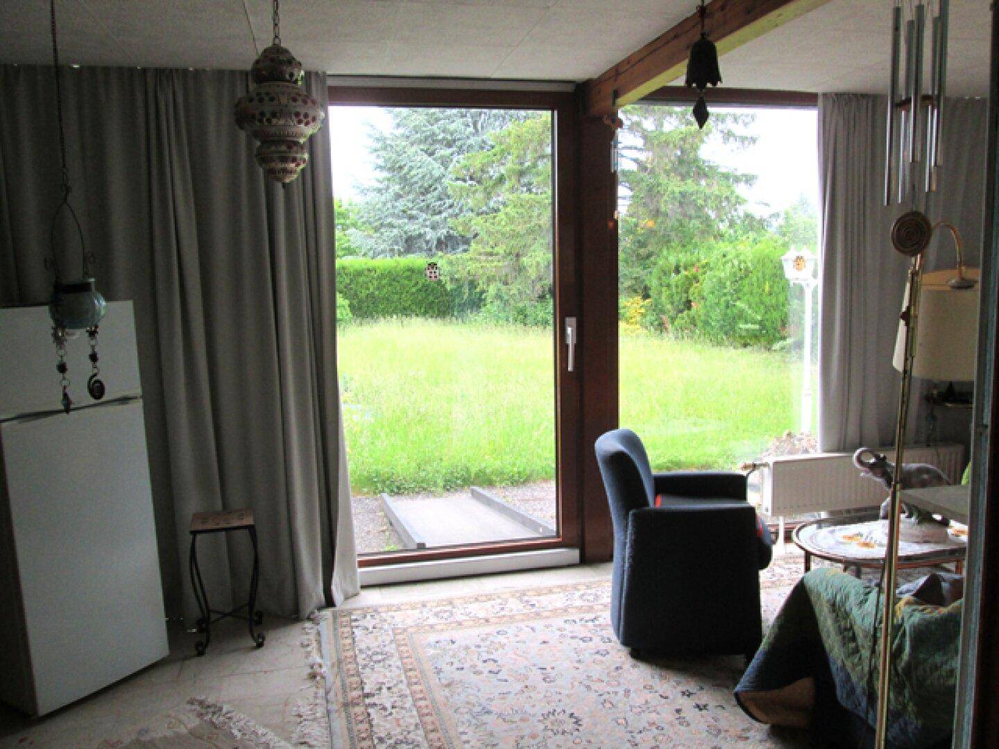Helles Wohnzimmer mit großen Fenstern, Blick ins Grüne und gemütlichem Sessel.