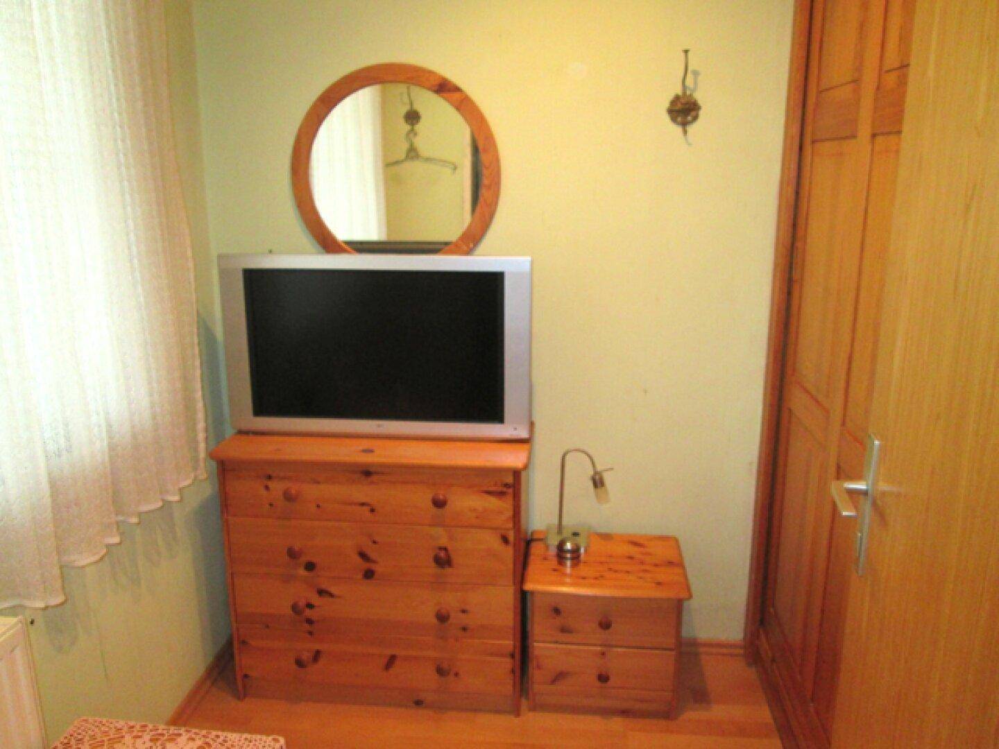 Schlafzimmer mit Holzkommode, Fernseher und schlichten Vorhängen.