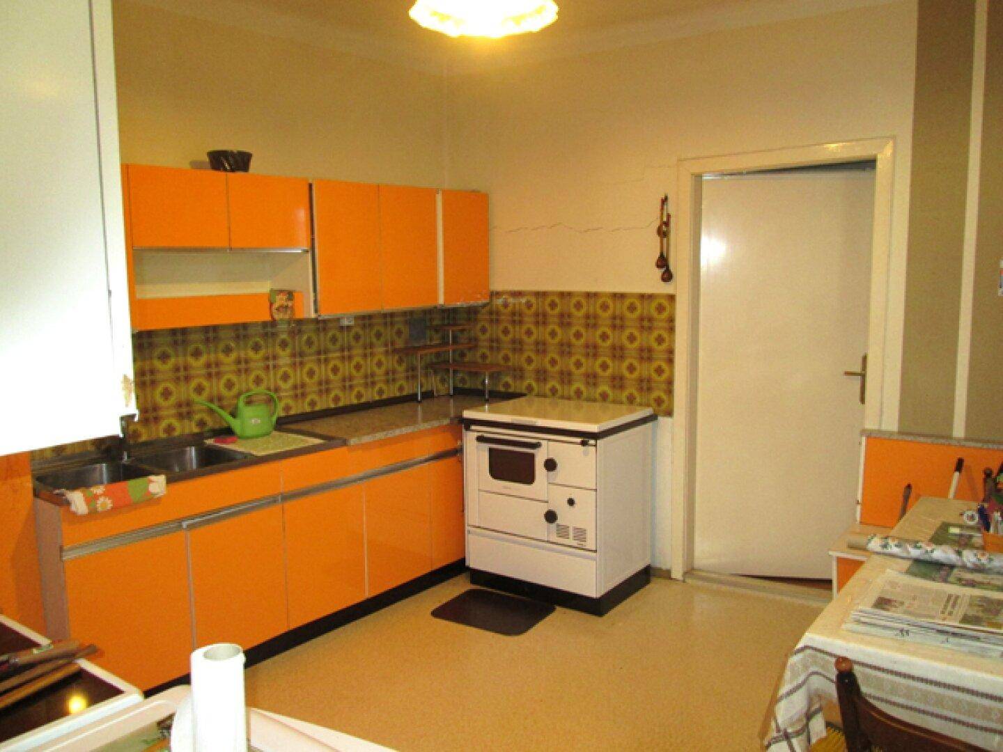 Küche im 80er-Jahre-Stil mit leuchtend orangefarbenen Schränken, gemusterten Fliesen und einem freistehenden Herd.