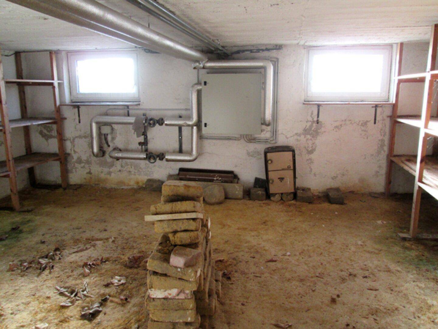 Unausgebauter Kellerraum mit sichtbaren Rohren, kleinen Fenstern und einfachen Holzregalen.