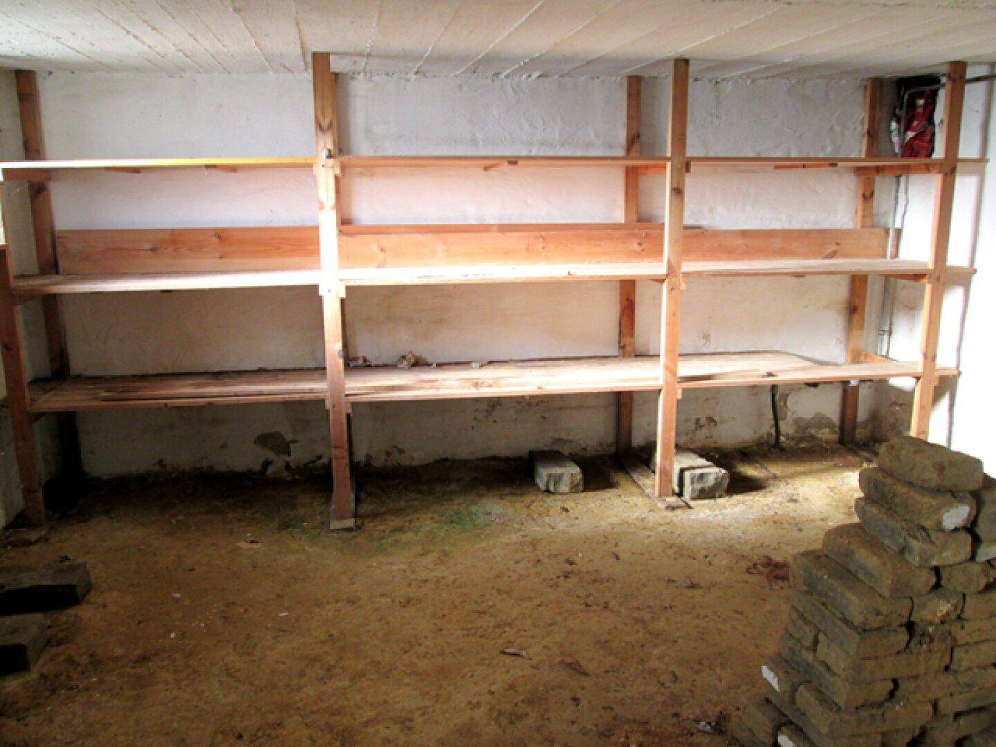 Kellerraum mit einfachen Holzregalen entlang der Wand und einem unfertigen Boden.