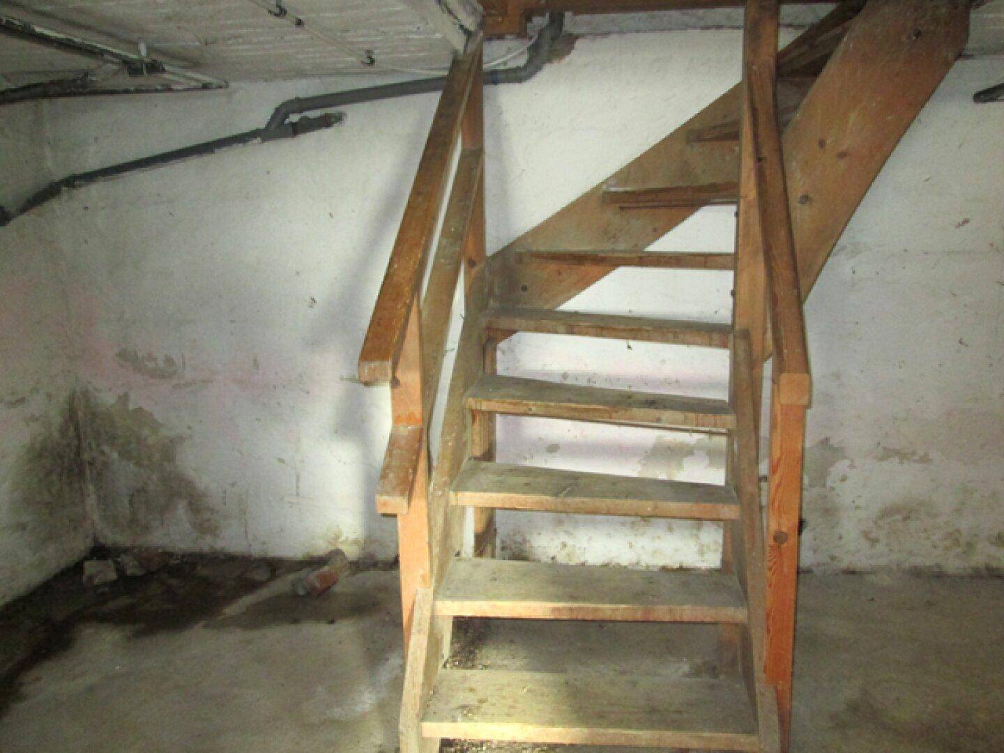 Kellerraum mit einer einfachen Holztreppe und sichtbaren Rohren an der Decke.