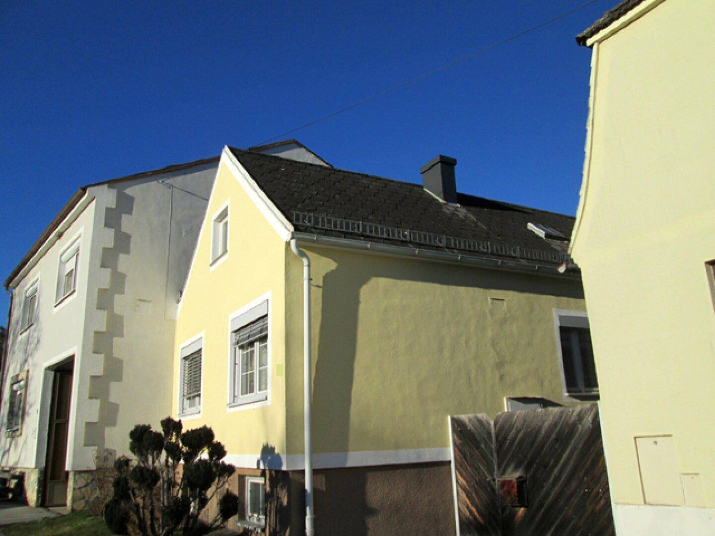 Seitenansicht des Hauses mit gelber Fassade, Giebeldach und Rollläden an den Fenstern.