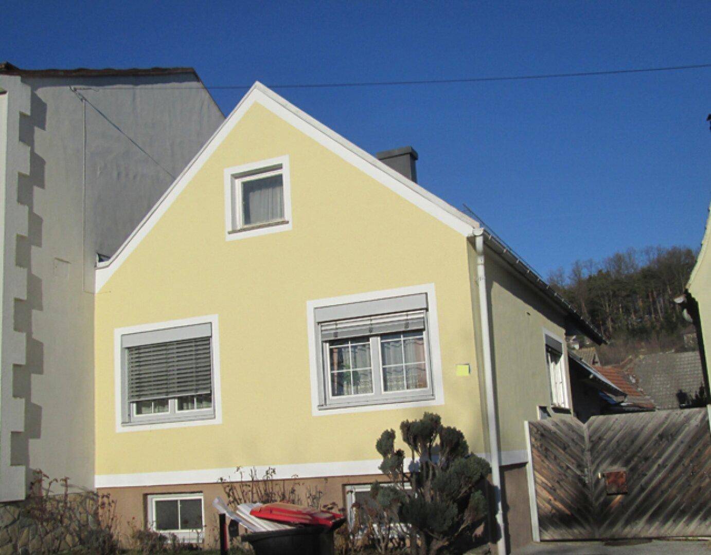 Vorderansicht des Hauses mit gelber Fassade, Giebeldach und Fenstern mit Rollläden.