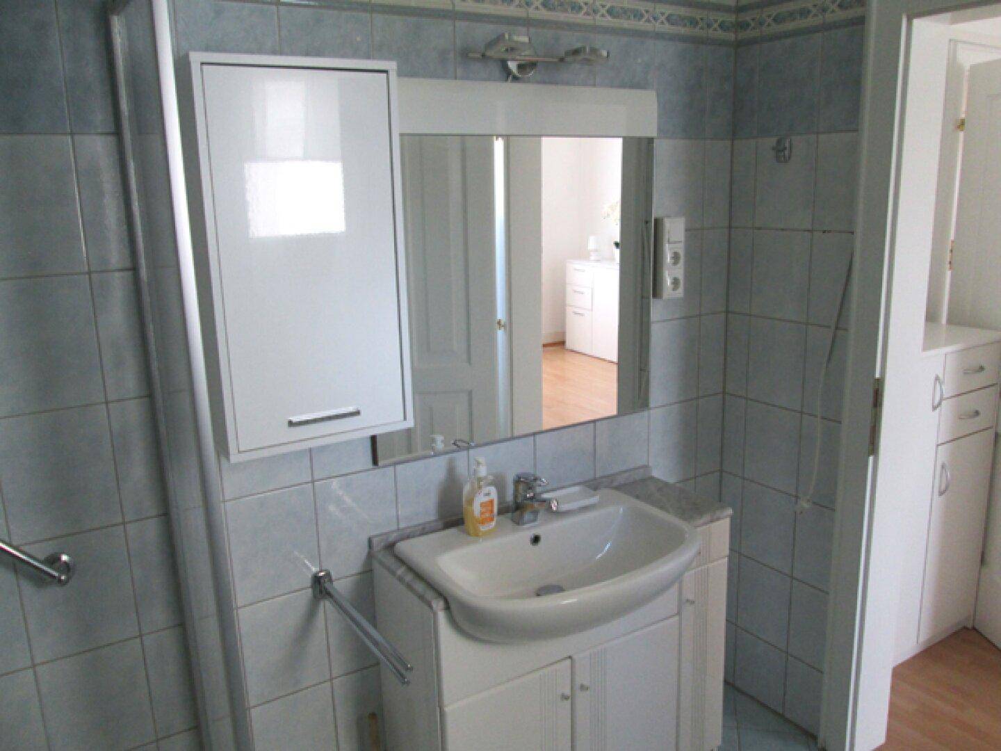 Badezimmer mit Waschbecken, Spiegelschrank und Duschbereich, gefliest in Blau- und Weißtönen.