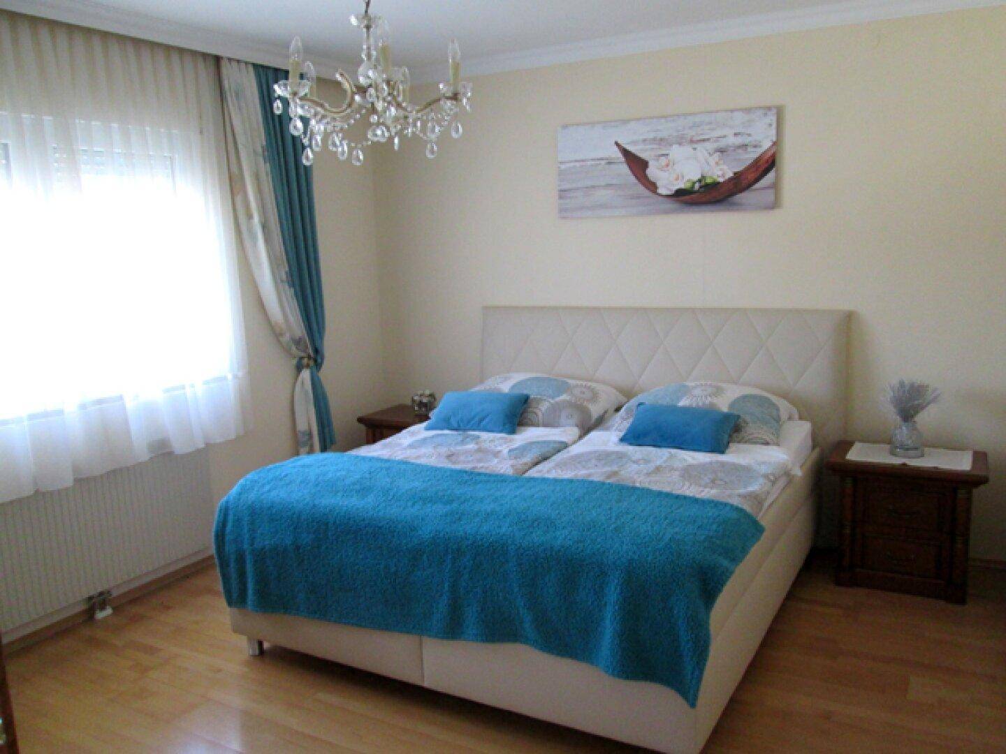Helles Schlafzimmer mit Doppelbett, großem Fenster und einem eleganten Kronleuchter an der Decke.