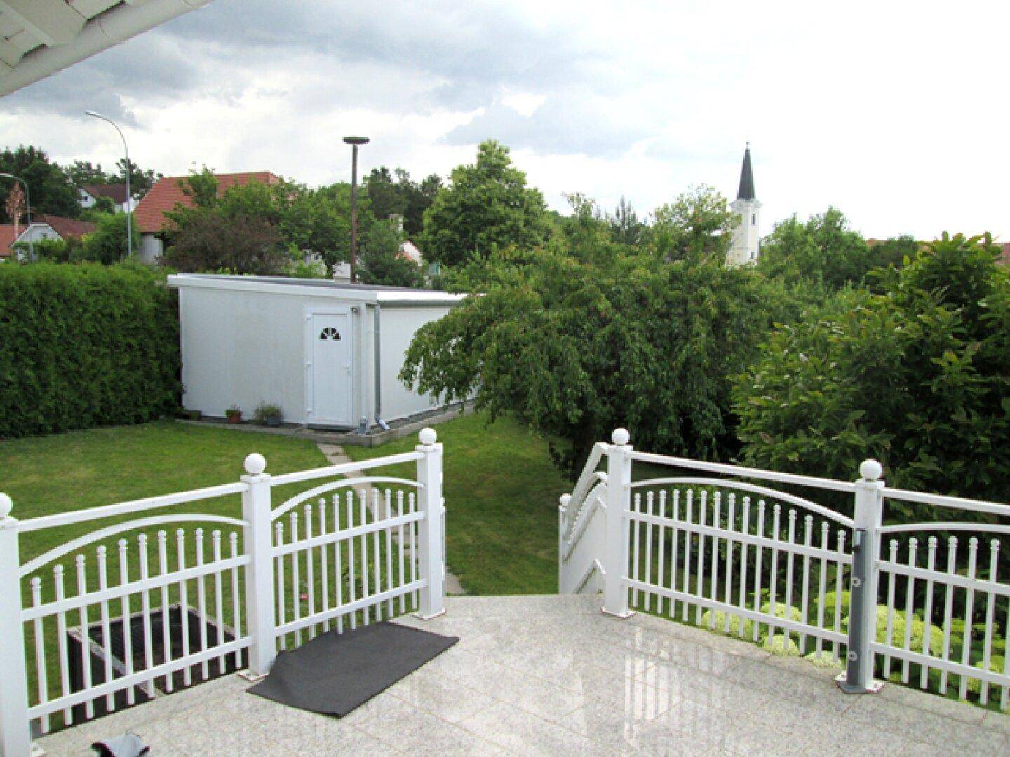 Terrasse mit Granitfliesen und weitem Blick über den grünen Garten bis zur Kirchturmspitze.