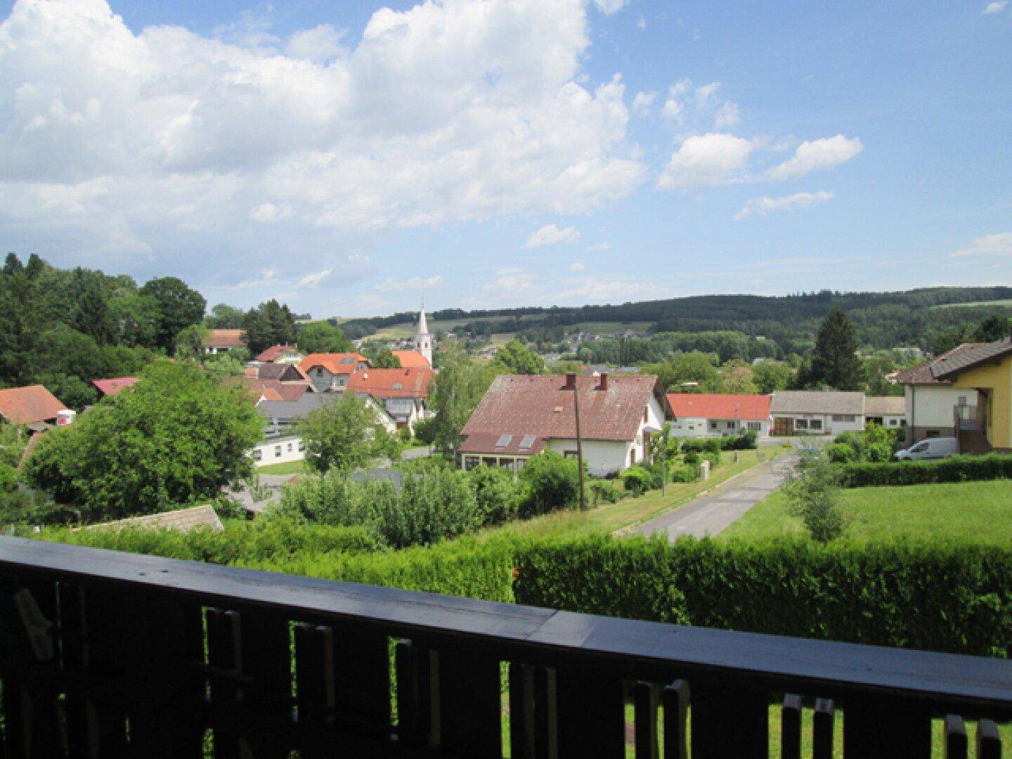 Weitläufiger Blick über eine ländliche Siedlung mit Häusern und grünen Hügeln von einem Balkon.