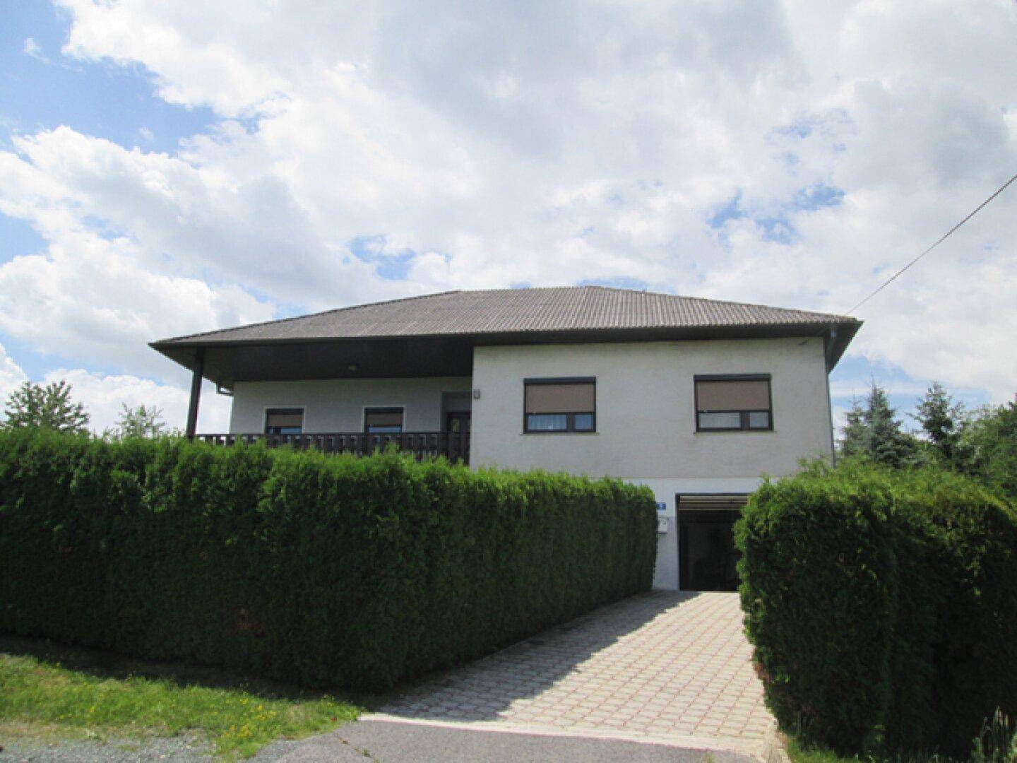 Vorderansicht des Hauses mit gepflasterter Auffahrt, grüner Hecke und Rollläden an den Fenstern.