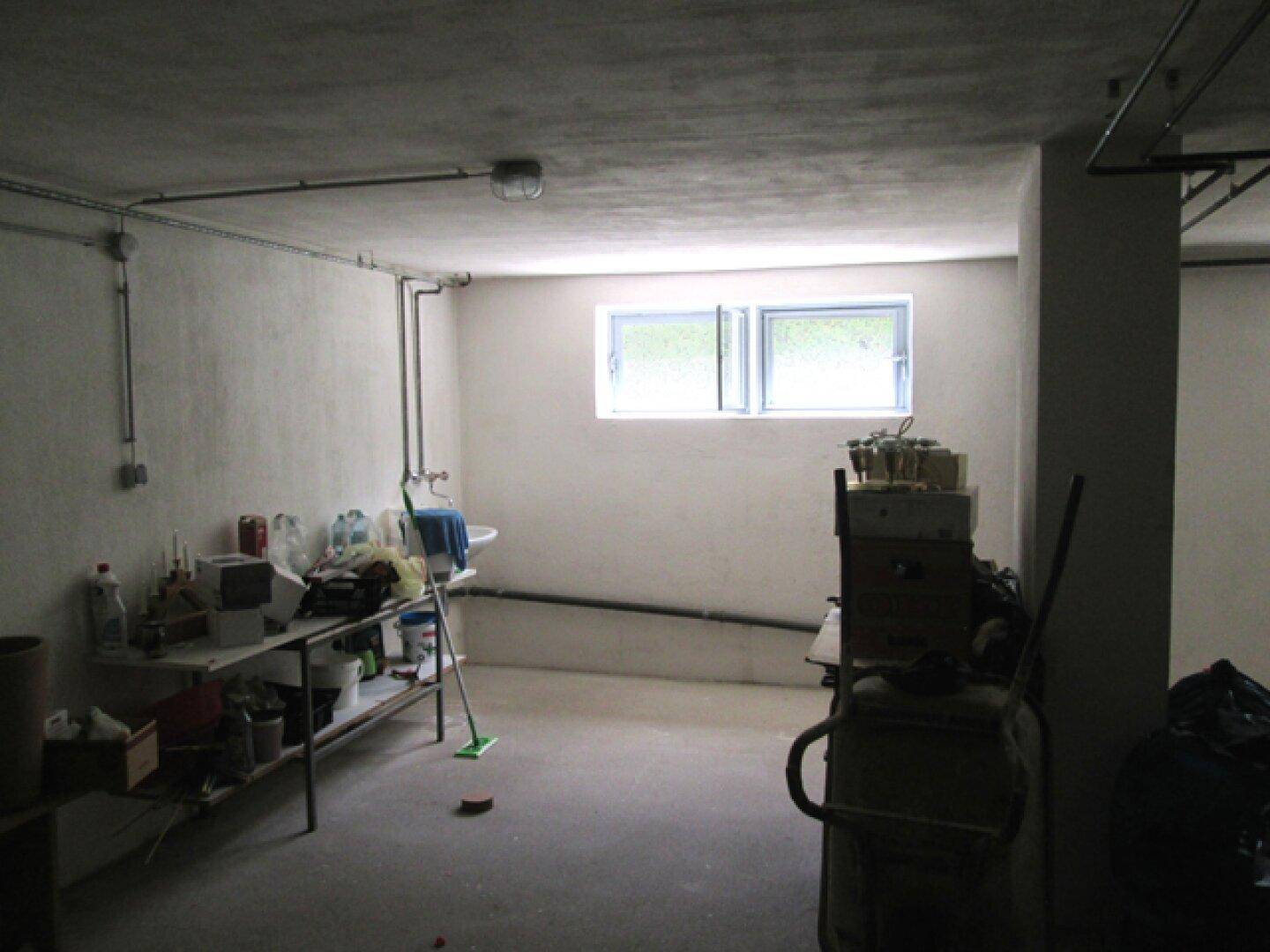 Unfertiger Kellerraum mit Betonboden, kleinem Waschbecken, sichtbaren Rohren und Fenster.