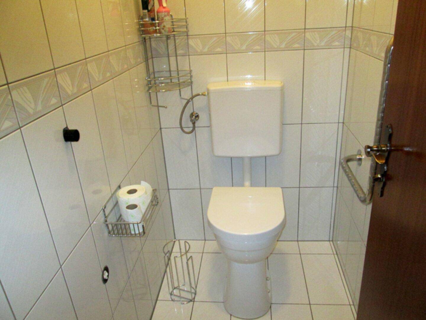 Separate Toilette mit weißen Fliesen, wandmontiertem WC und kleinem Regal für Toilettenpapier.