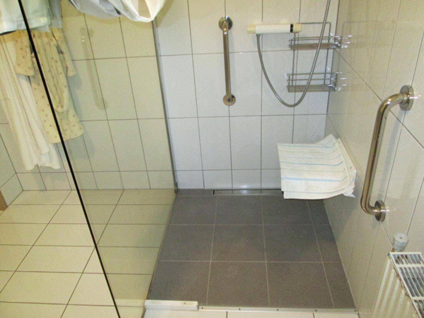 Duschbereich mit weißen und grauen Fliesen, Glasabtrennung und Haltegriffen für Sicherheit.
