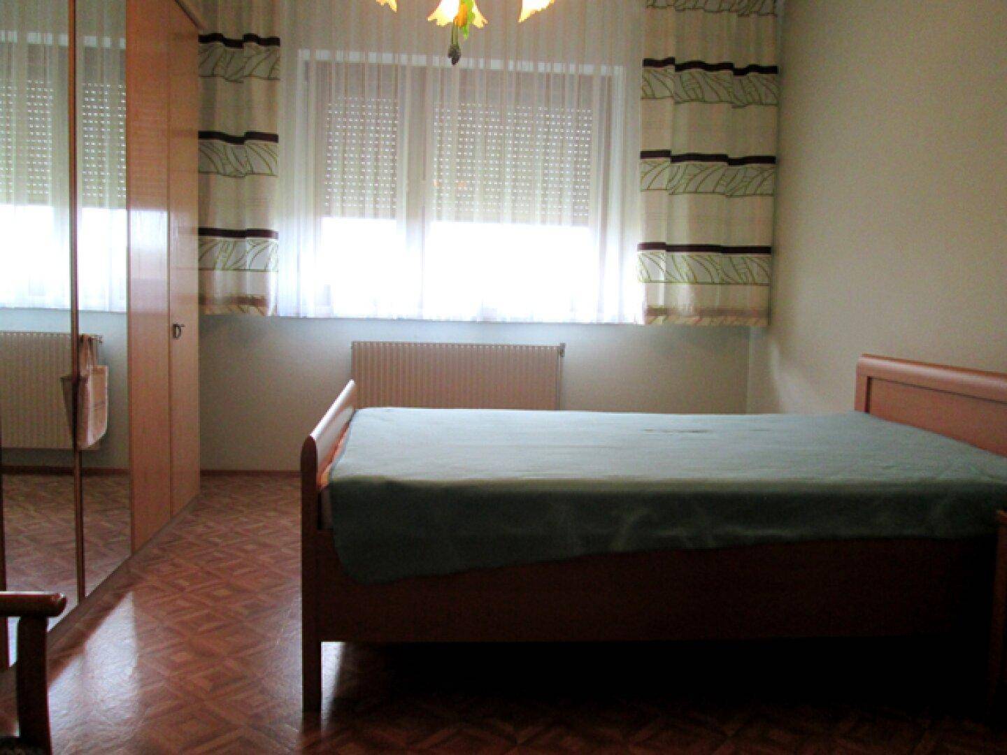 Schlafzimmer mit Holzbett, Spiegelschrank und Fenster mit Vorhängen für Privatsphäre.