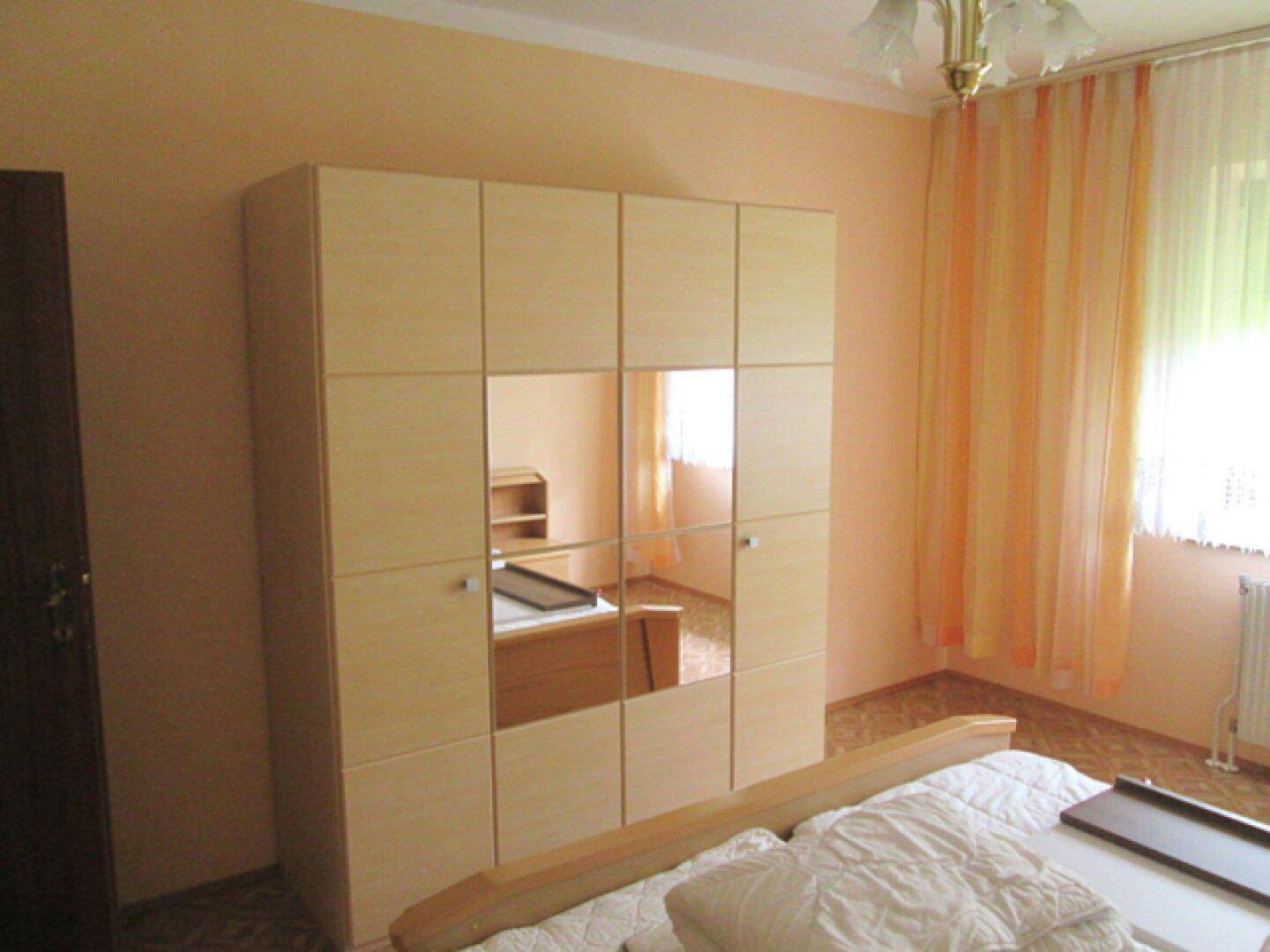 Helles Schlafzimmer mit großem Spiegelschrank, Bett und Fenster mit orangefarbenen Vorhängen.